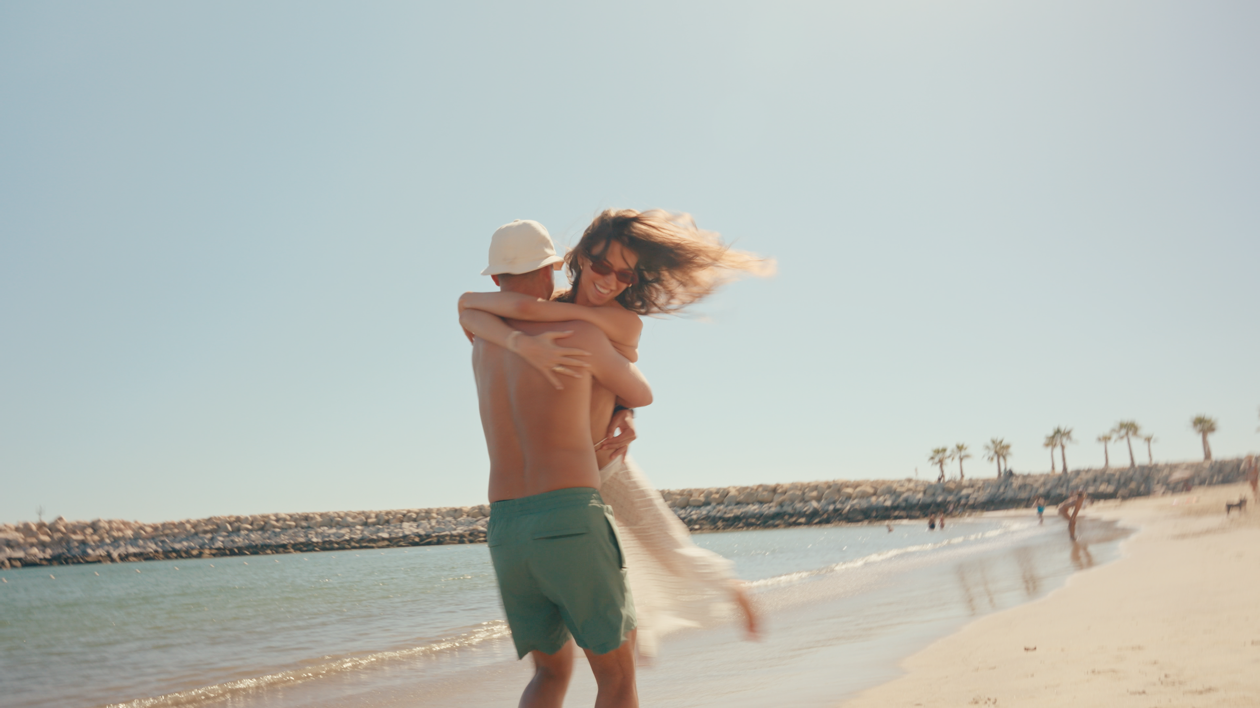 A man lifting a woman on the beach, both smiling and embracing, with palm trees and the ocean in the background.
