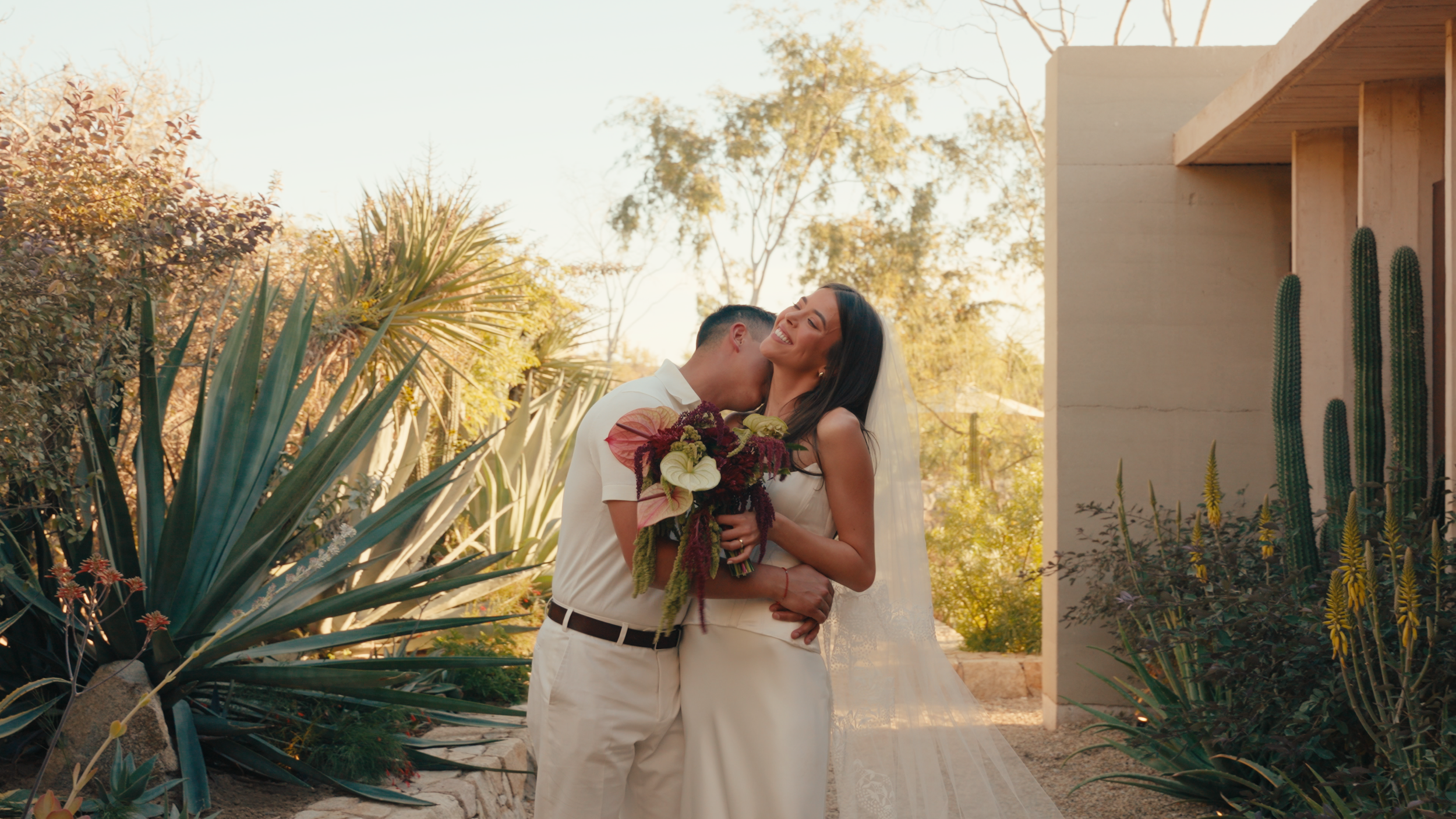 A bride and groom share a moment outdoors, with the groom kissing the bride on the cheek. The bride is smiling, holding a bouquet of flowers, and has long dark hair, while the groom is wearing a white shirt and light-colored trousers. The scene is su