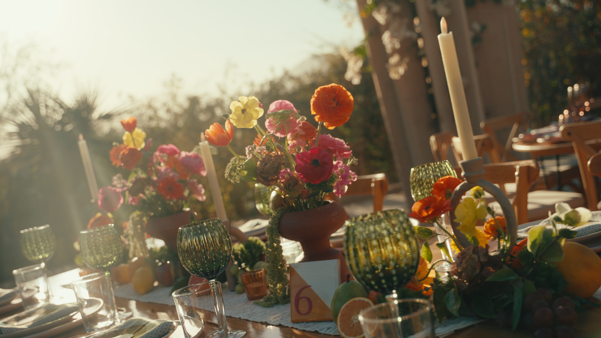 Elegant outdoor dining table decorated with floral centerpieces, candles, green glassware, and fruit, illuminated by warm sunlight.