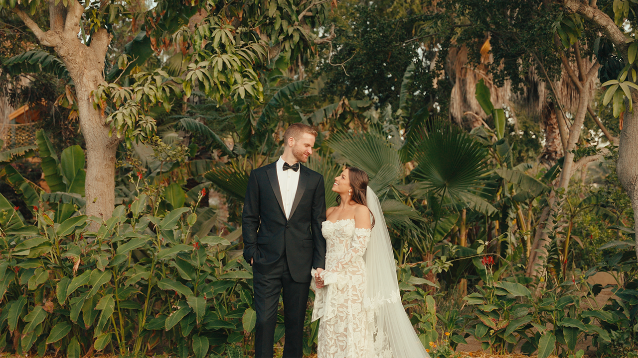 A bride and groom holding hands, standing in a lush, green garden with large leaves and trees, facing each other and smiling.