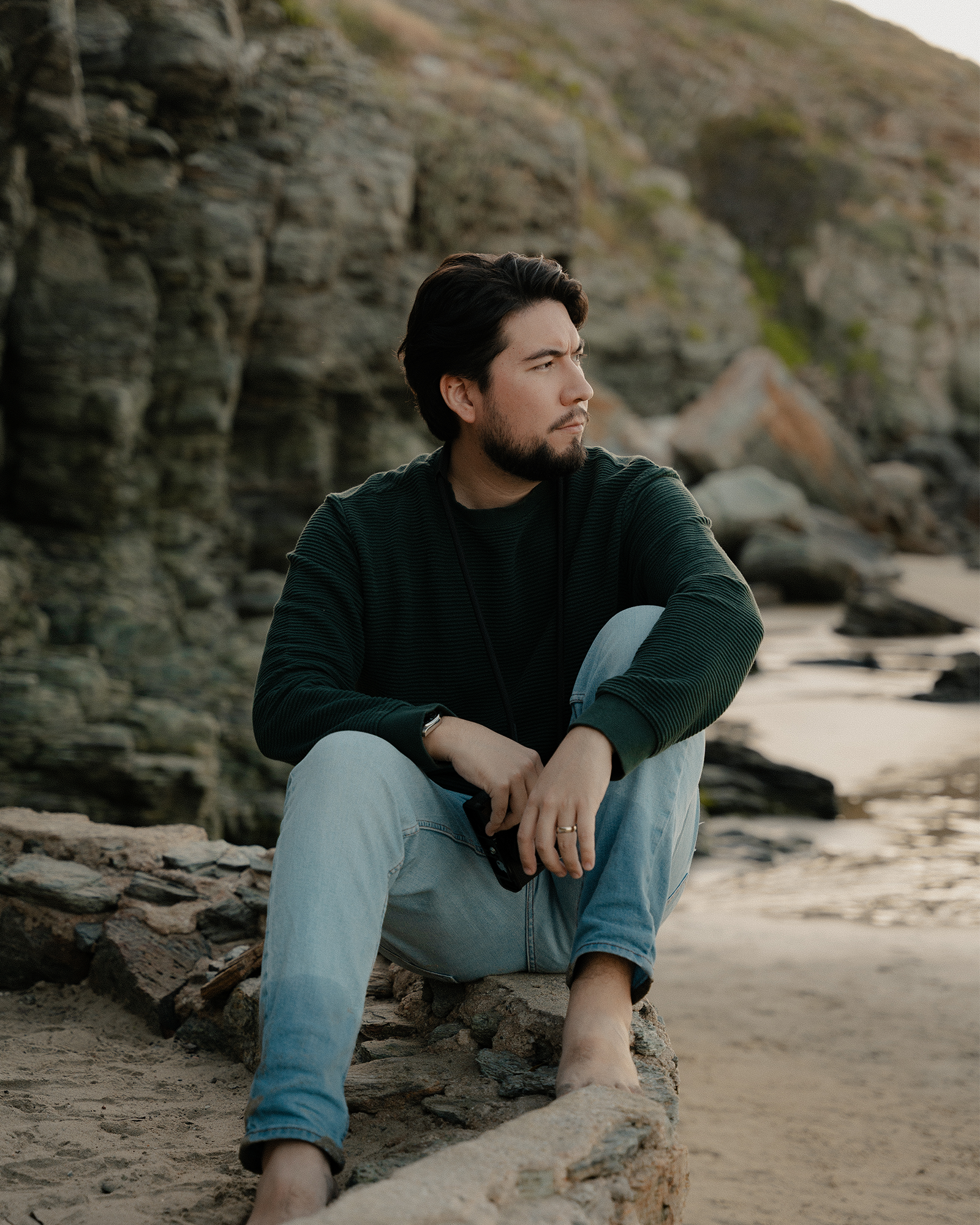 A man with dark hair and beard sitting on a rocky beach near cliffs, looking pensively into the distance, wearing a dark long-sleeve shirt and light jeans.