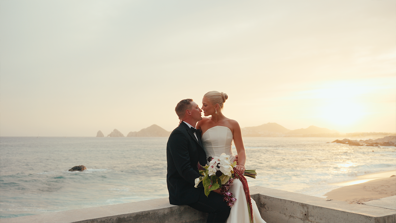 A bride and groom sitting close on a concrete ledge by the sea during sunset, the bride holding a bouquet of white and purple flowers.
