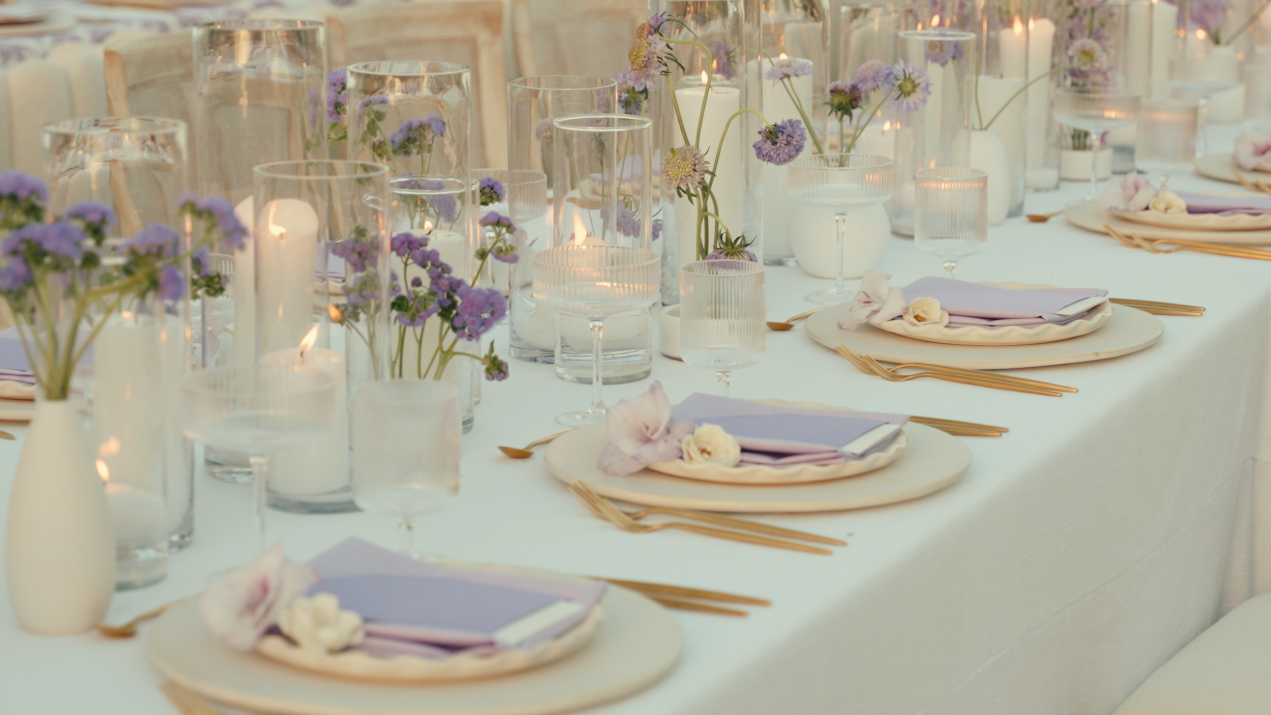 Elegant table setup for a wedding or special event with white tablecloth, lavender flowers, candles, and neatly arranged place settings with plates, napkins, and gold utensils.
