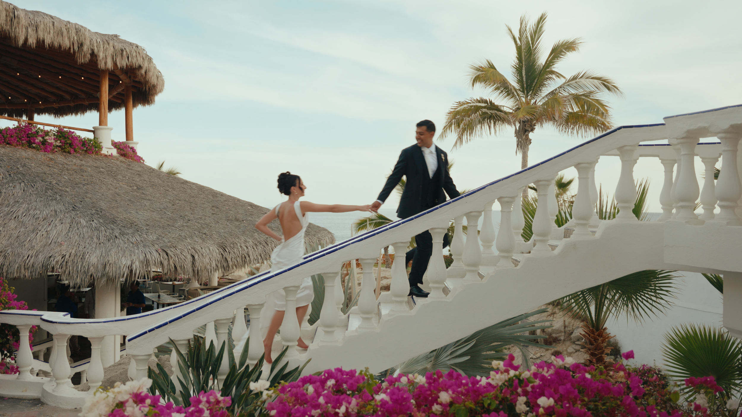 A couple in wedding attire holding hands on a staircase at a beachside resort with palm trees, pink and purple flowers, and thatched-roof buildings.