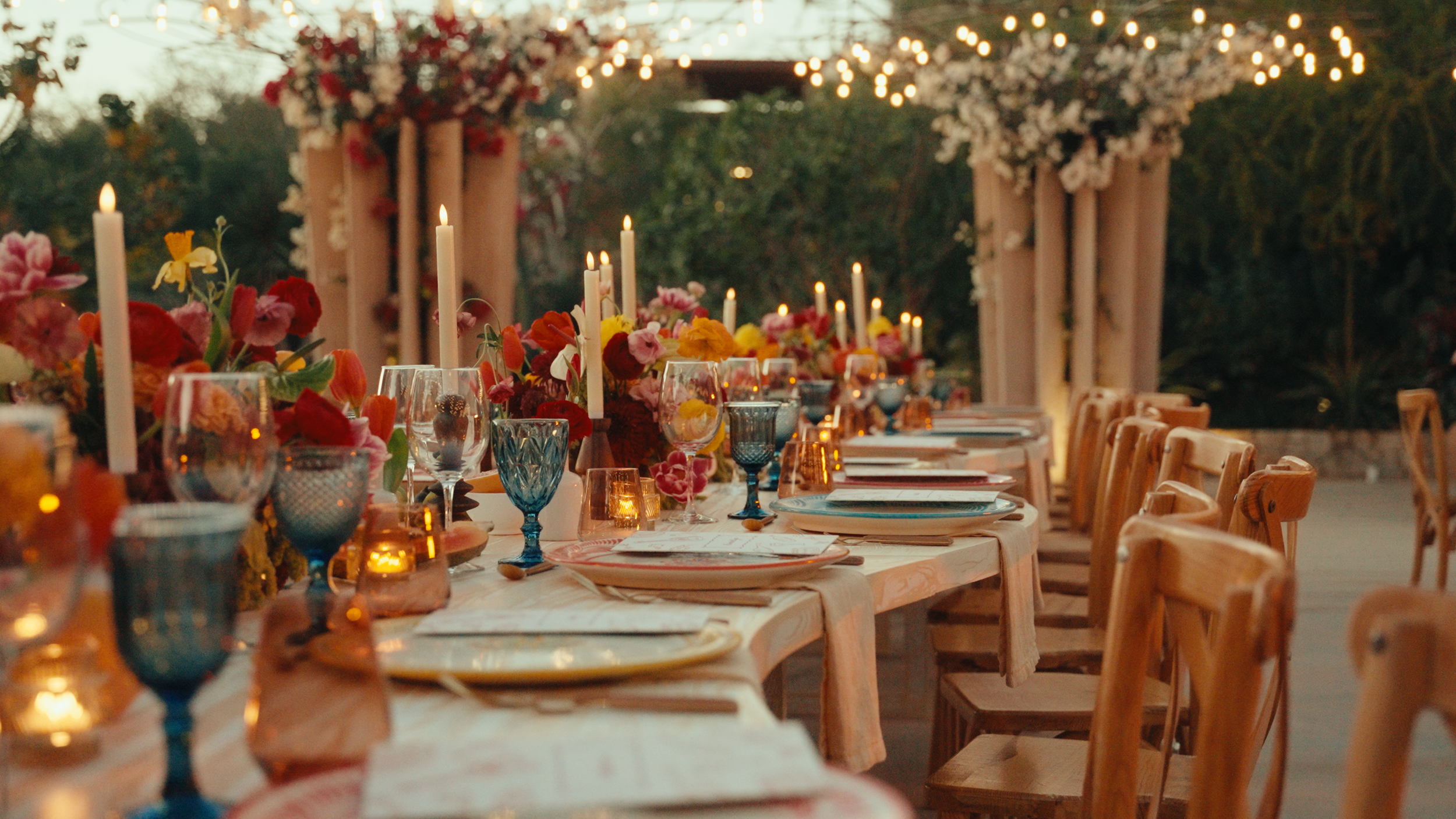 Elegant outdoor dinner table decorated with flowers, candles, and glassware under string lights at sunset.