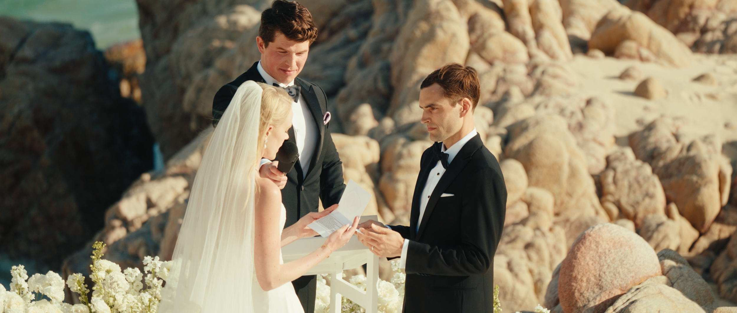 A wedding ceremony taking place outdoors on a rocky beach with three people—bride, groom, and officiant—standing together. The bride wears a white dress and veil, reading vows or a speech. The groom is dressed in a black tuxedo and holds her hand. Th