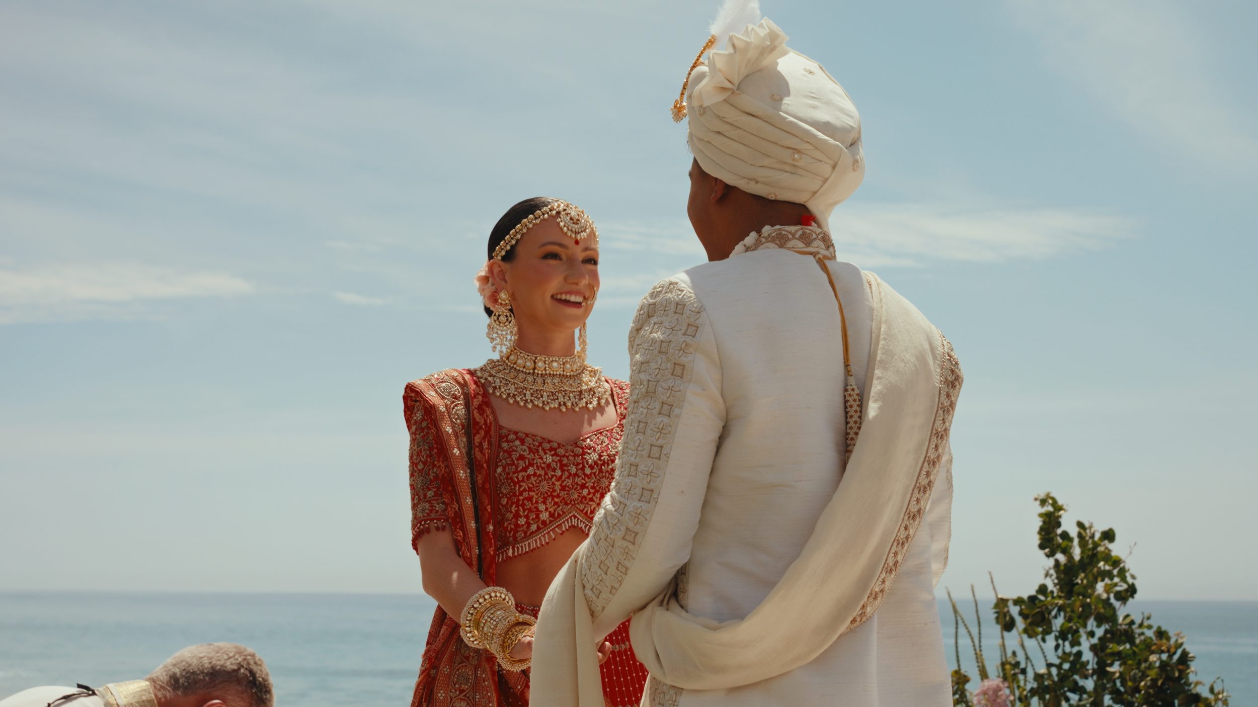 A couple in traditional Indian wedding attire standing outdoors near the ocean, exchanging vows, with clear sky and some plants in the background.