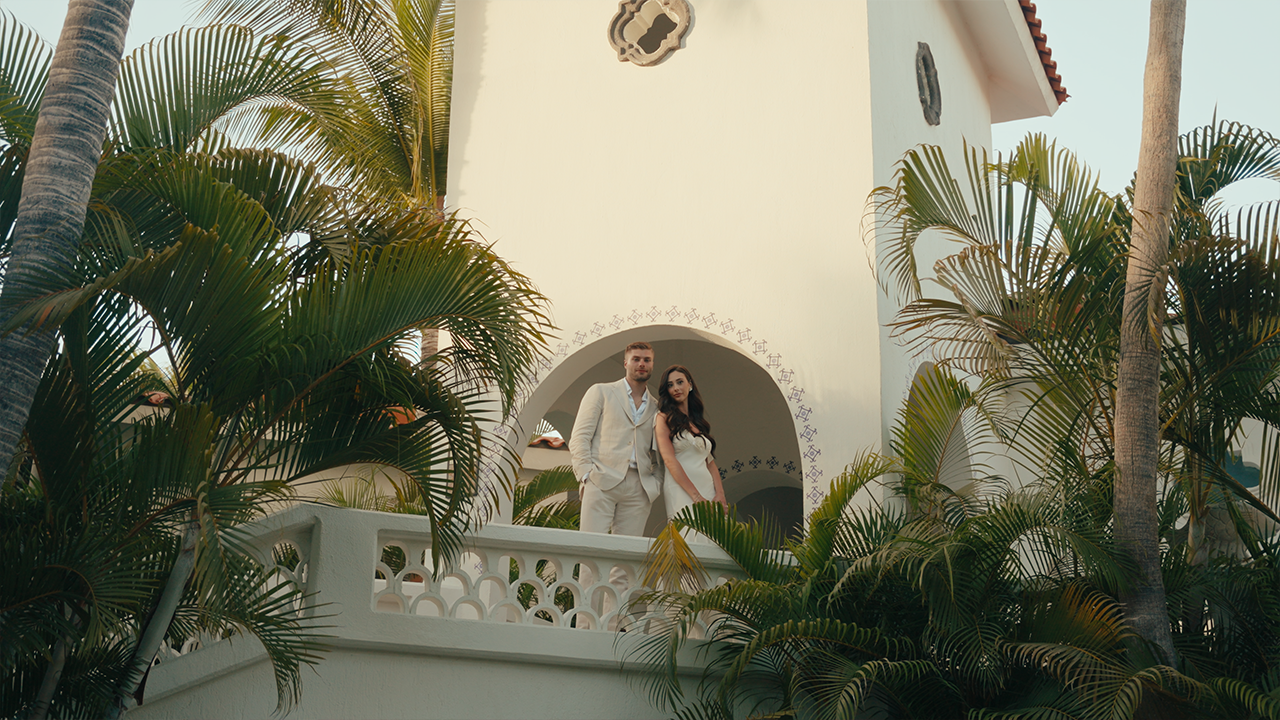 A couple dressed in white clothing standing on a balcony of a white building surrounded by tropical plants like palm trees.
