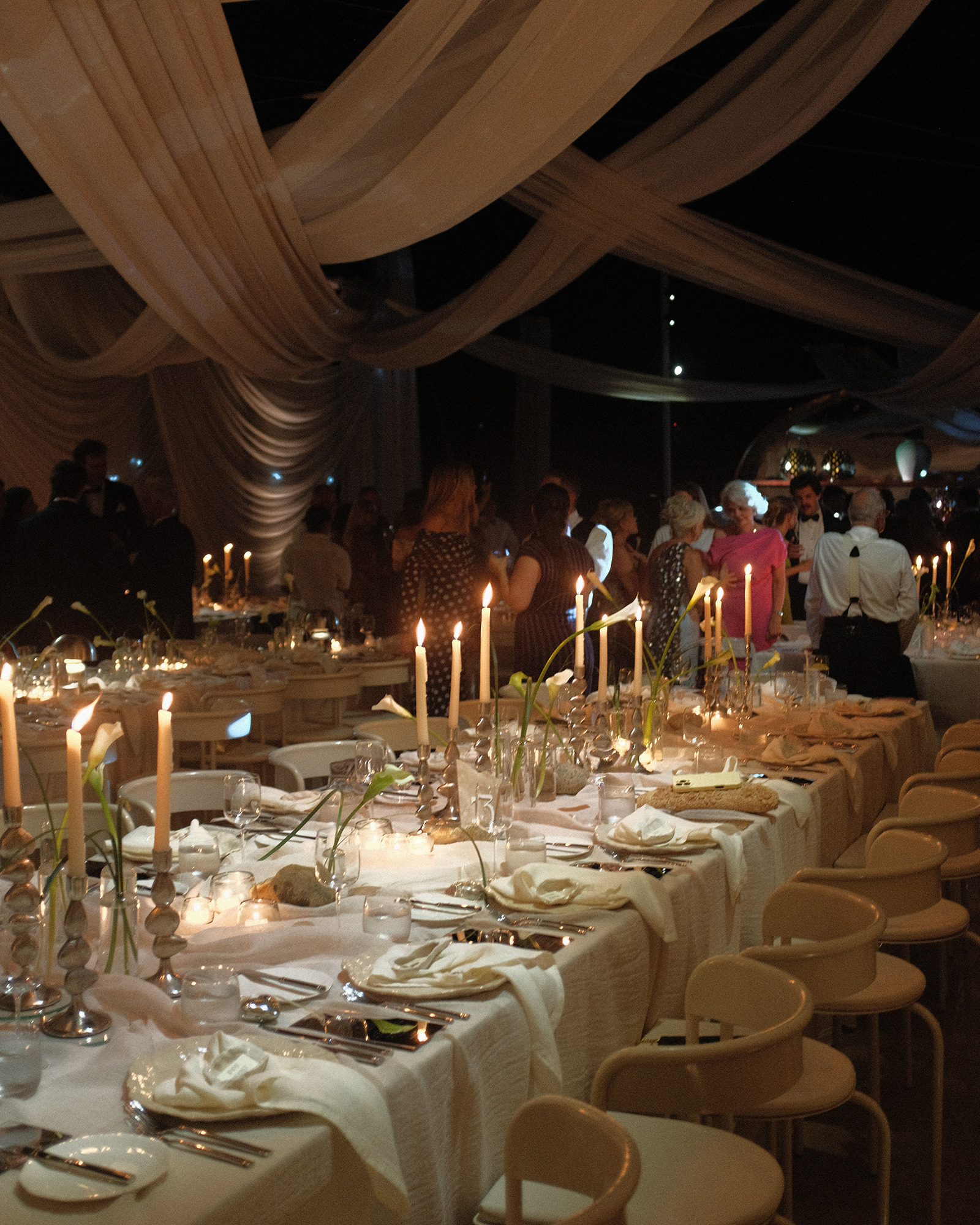 Elegant banquet hall decorated with hanging fabric drapes, candlelit tables, and guests dressed in formal attire mingling.