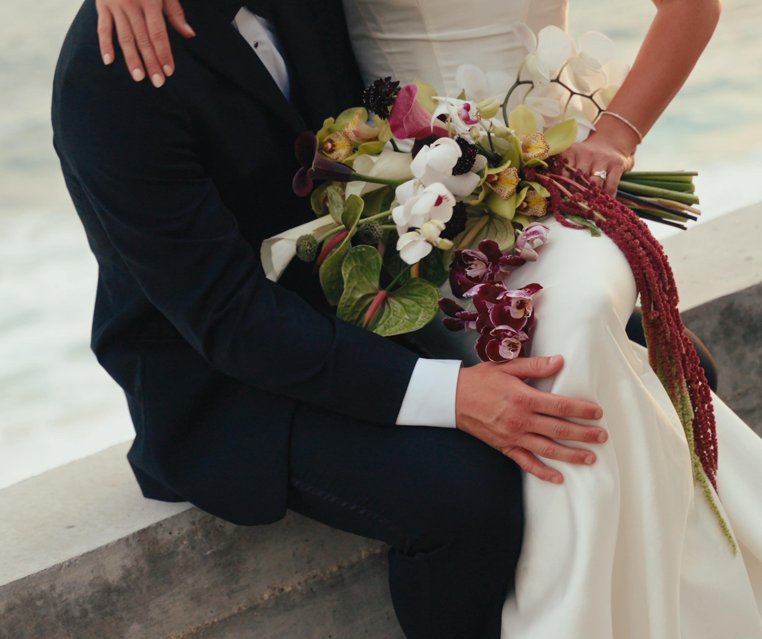 A bride and groom sitting by the water, with the bride holding a large bouquet of orchids and other flowers, wearing a white wedding dress, and the groom in a black tuxedo.