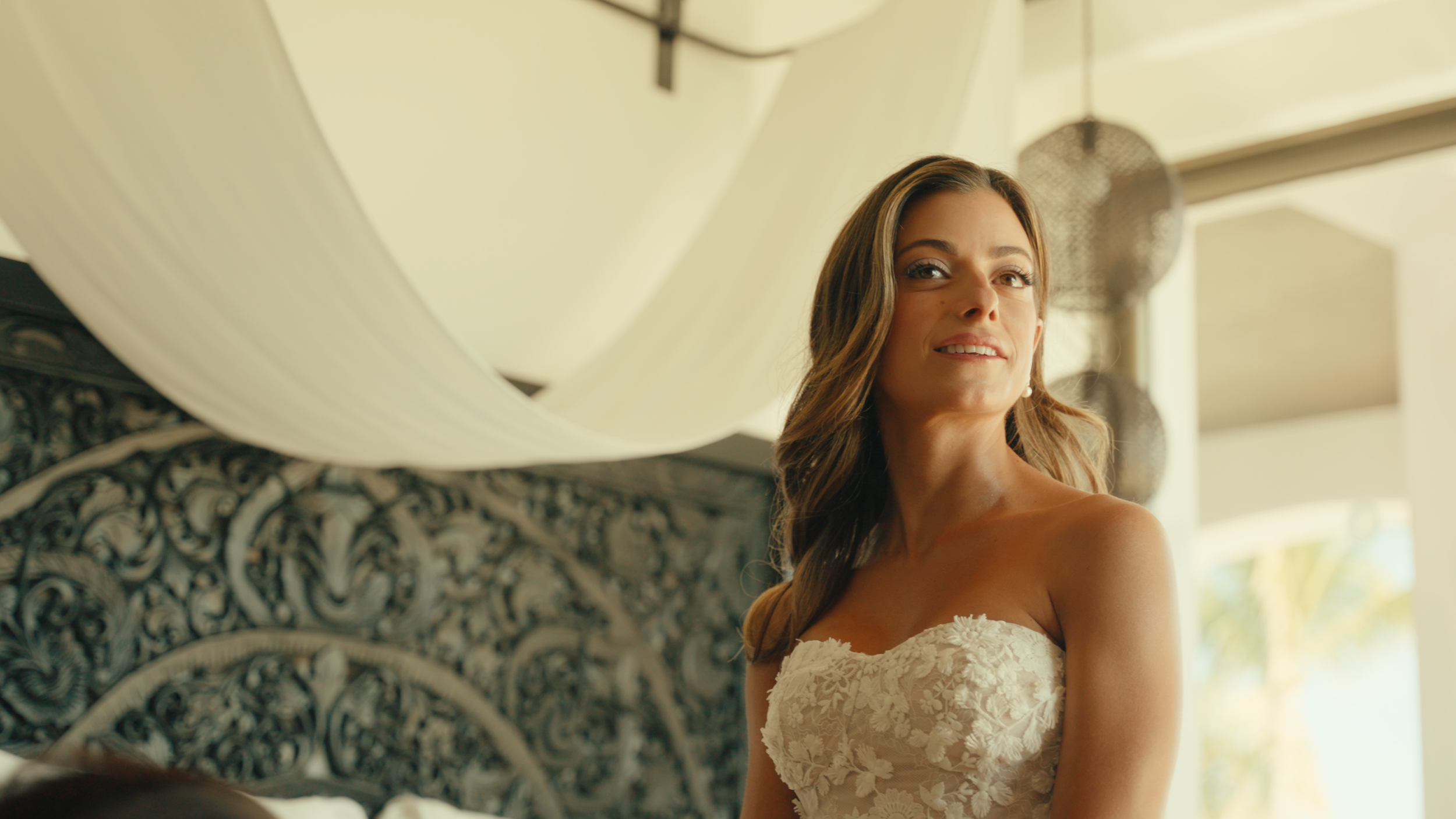 A woman in a wedding dress with lace details smiling and looking away, inside a well-lit room with hanging lanterns and large windows.