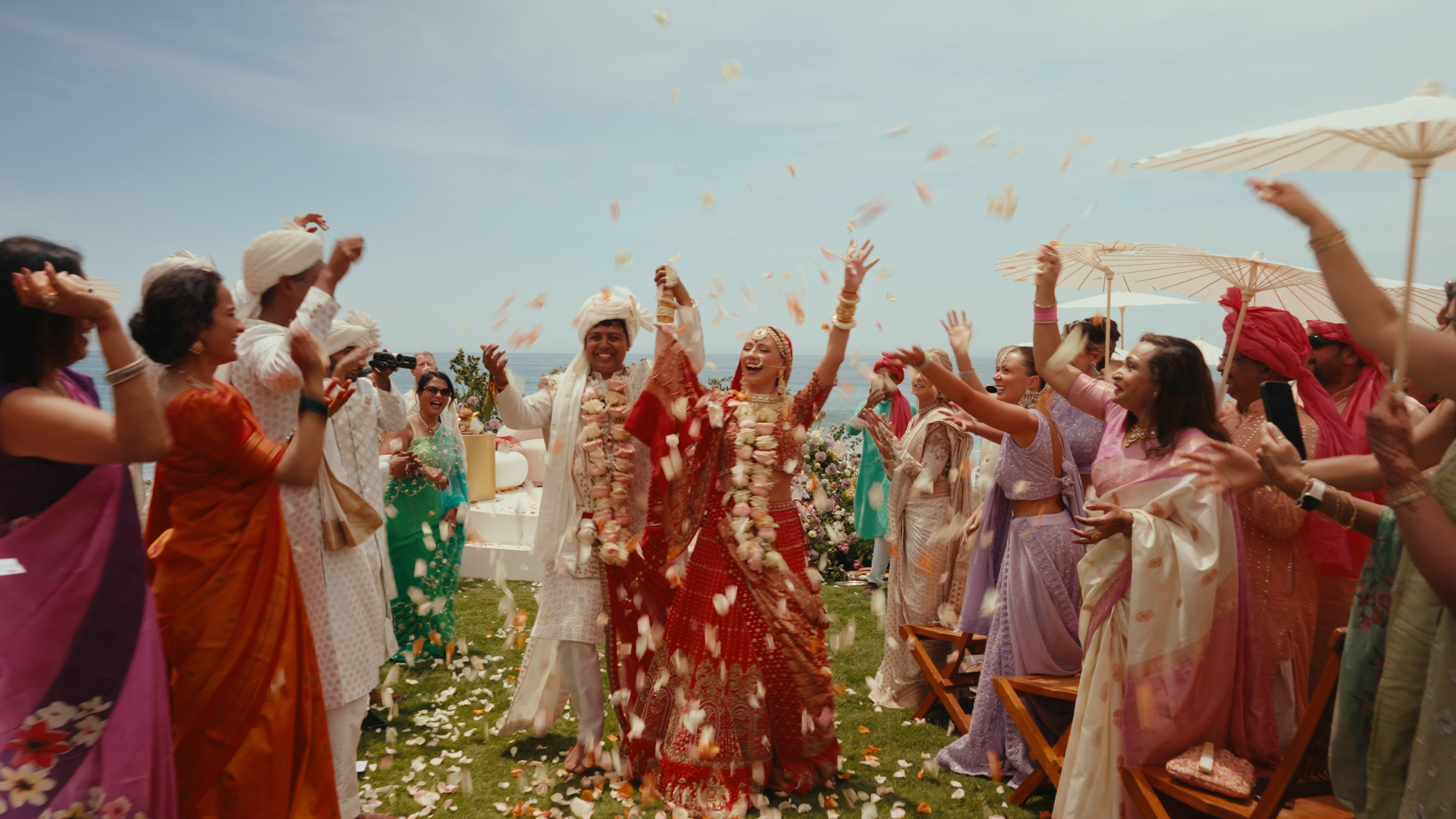 Indian wedding celebration outdoors with guests throwing flower petals at the bride and groom, who are dressed in traditional attire, with umbrellas and the ocean in the background.