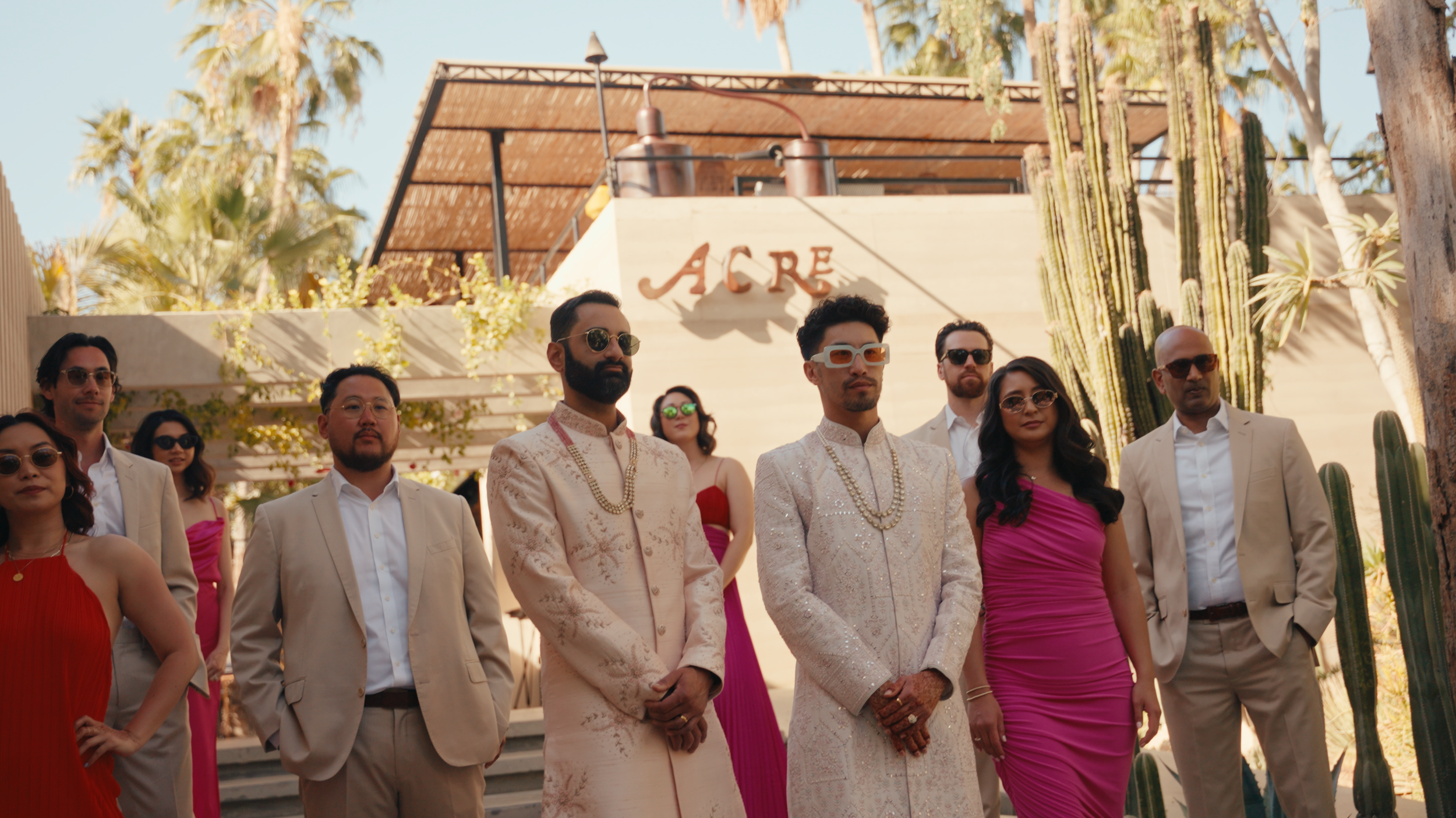 A group of men and women dressed in formal and colorful attire standing outdoors in front of a building with the word 'ACRE' on it, surrounded by palm trees and cacti.