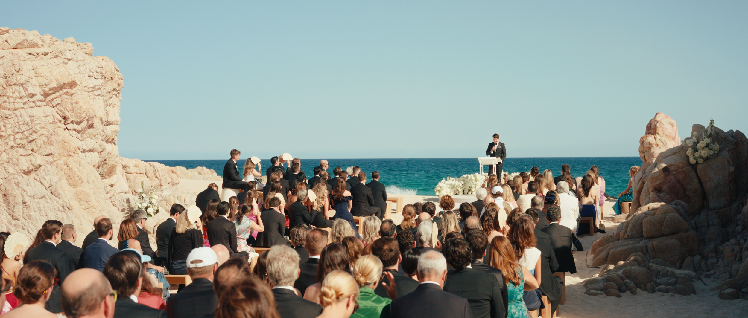 Wedding ceremony on a beach with a large crowd of guests seated in front of the ocean, with a man in a suit giving a speech at a podium decorated with white flowers.
