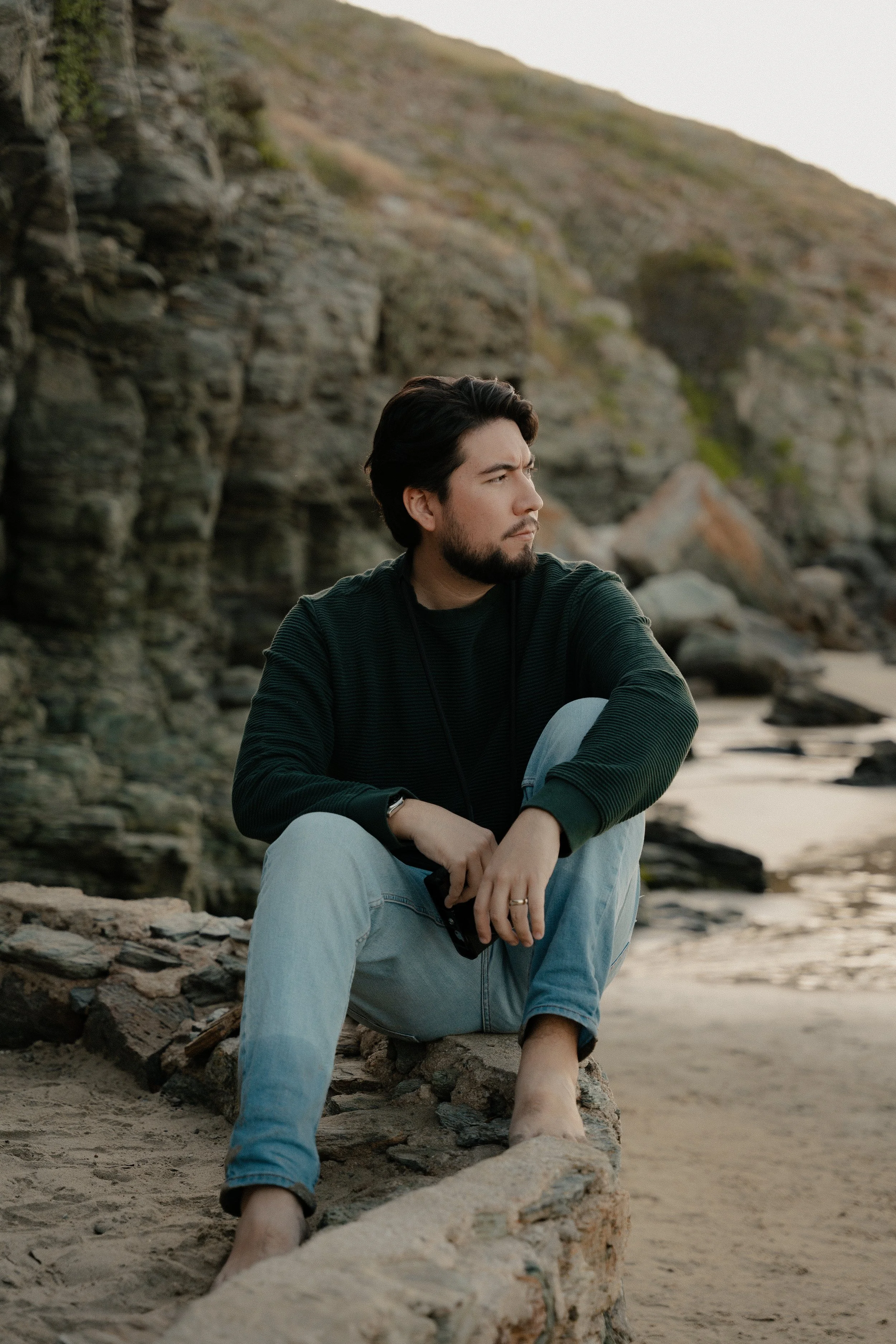 A man sitting on a rock at a beach during sunset, looking contemplative and gazing into the distance with rocky cliffs behind him.