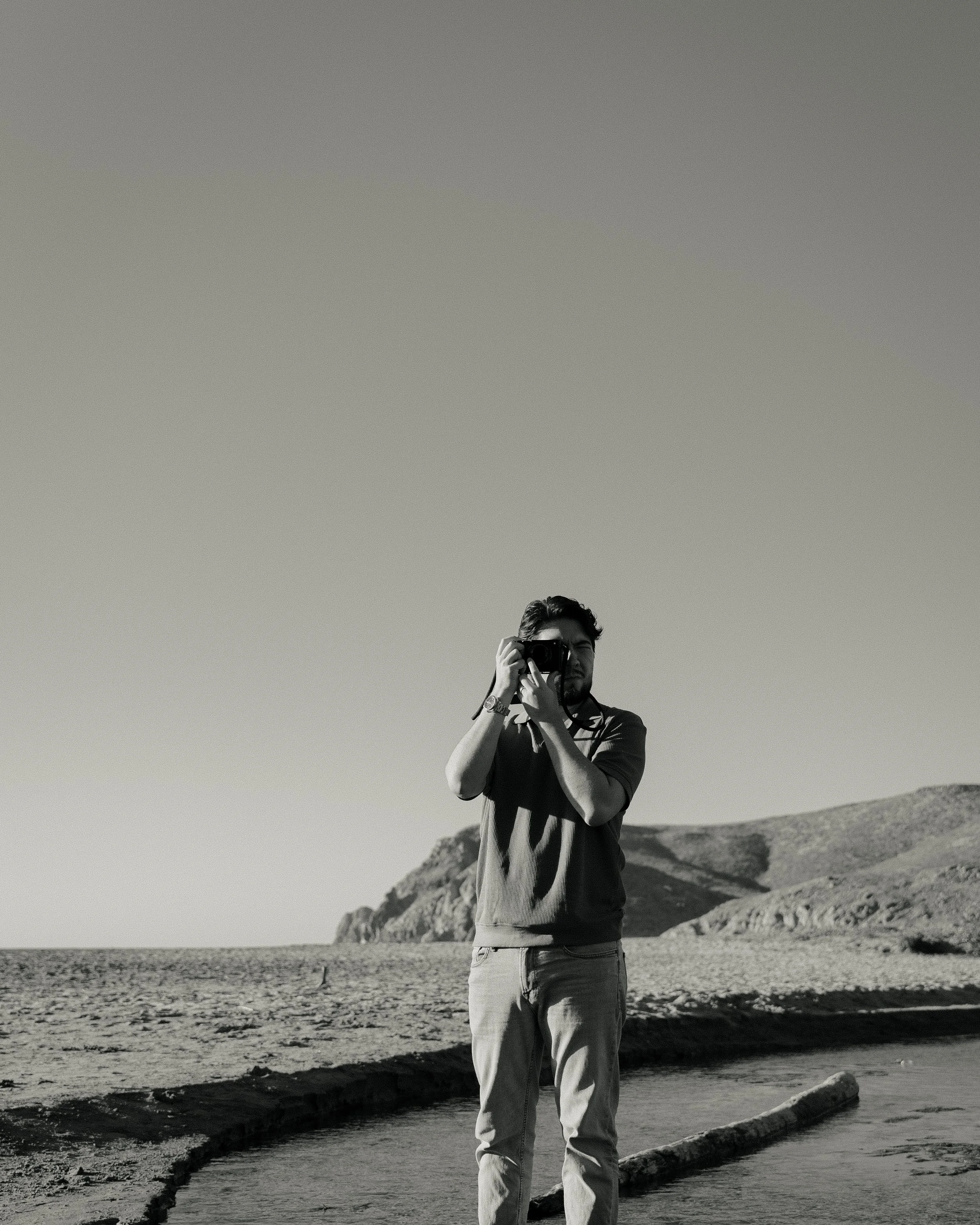 A man taking a photograph on a beach with hills in the background, in black and white.