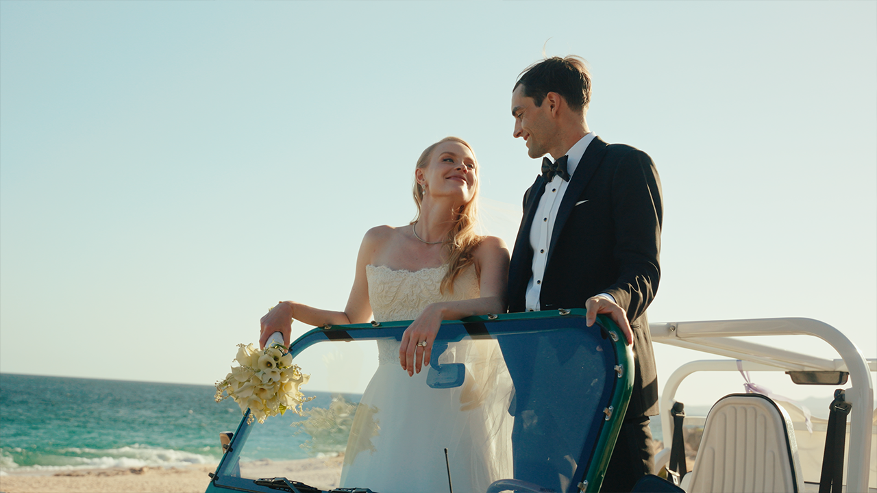 A bride and groom in wedding attire standing on a beach, looking at each other and smiling during sunset.