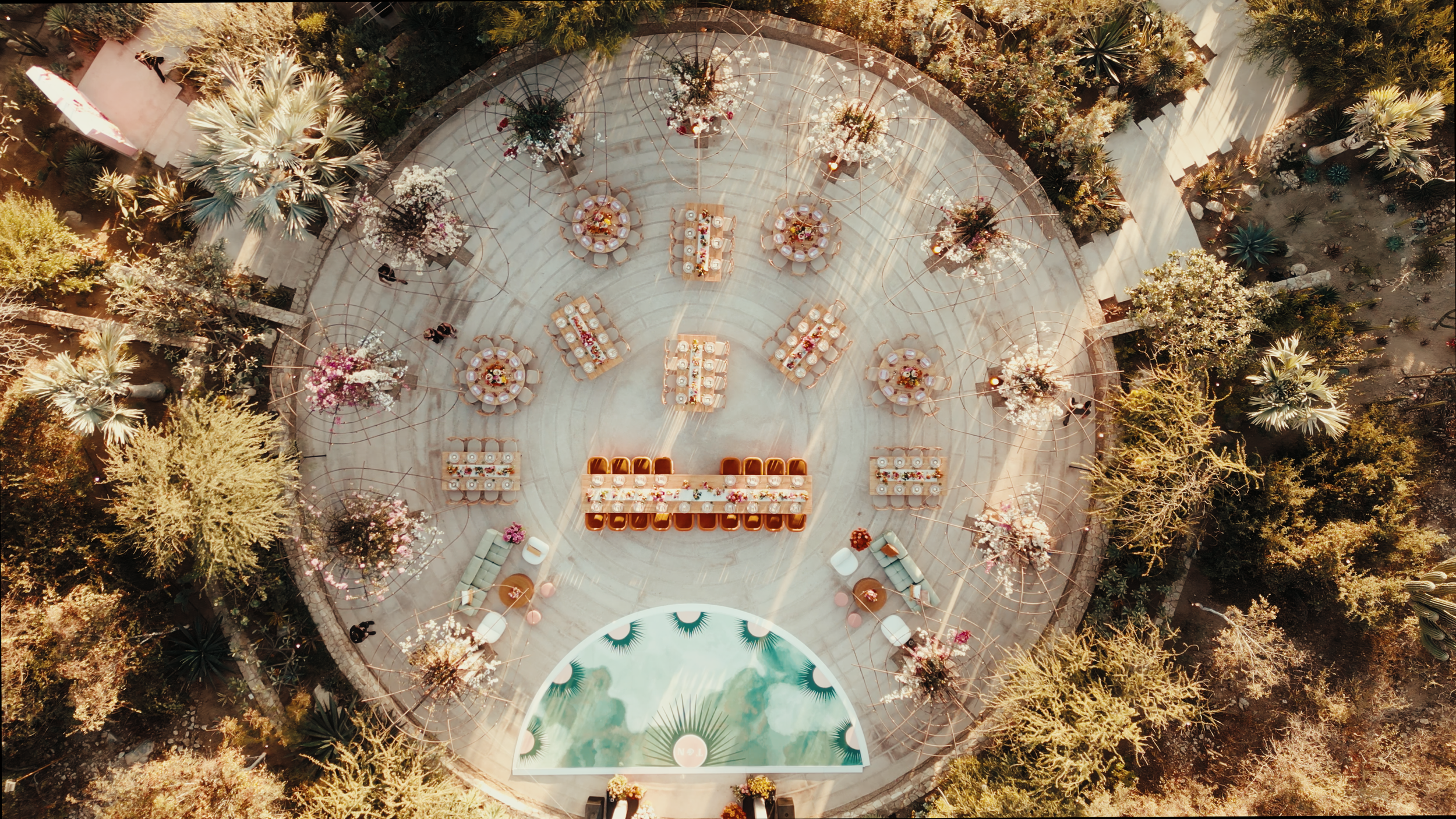 Aerial view of an outdoor event setup with round and rectangular tables decorated with flowers on a circular stone surface, surrounded by trees and desert plants.