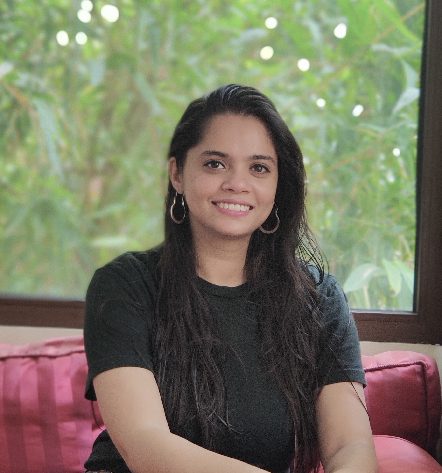 A young woman with long dark hair, wearing hoop earrings and a black shirt, sitting on a pink cushioned bench in front of a window with a green leafy background.