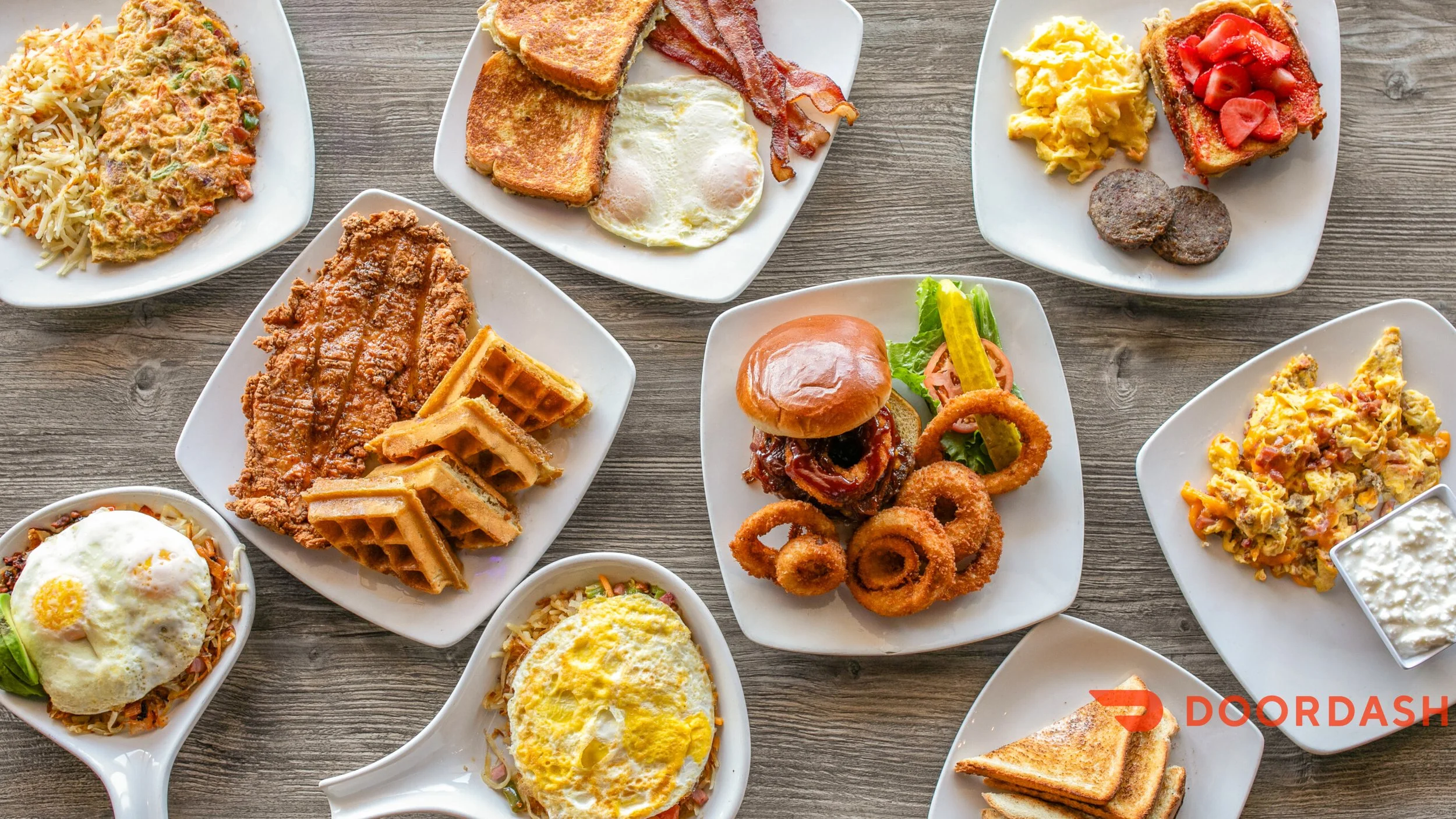 Assorted breakfast dishes including fried eggs, waffles, French toast, breakfast sandwich, scrambled eggs, fried chicken, biscuits and gravy, breakfast casserole, and sides of fruit and bacon, all on white plates on a wooden table.