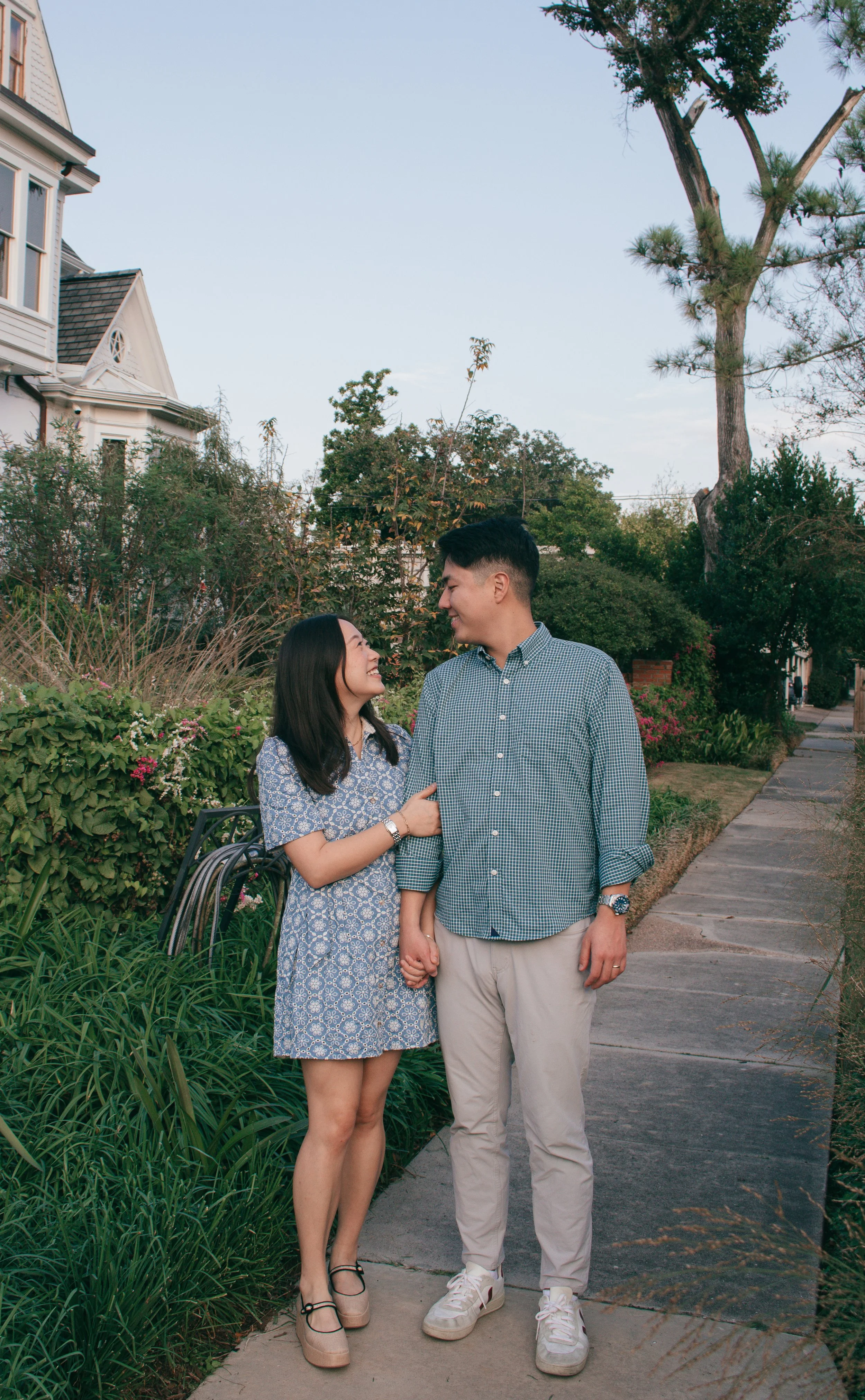 Lifestyle photo of couple standing hand in hand outdoors, framed by lush plants, a white house, and warm daylight.