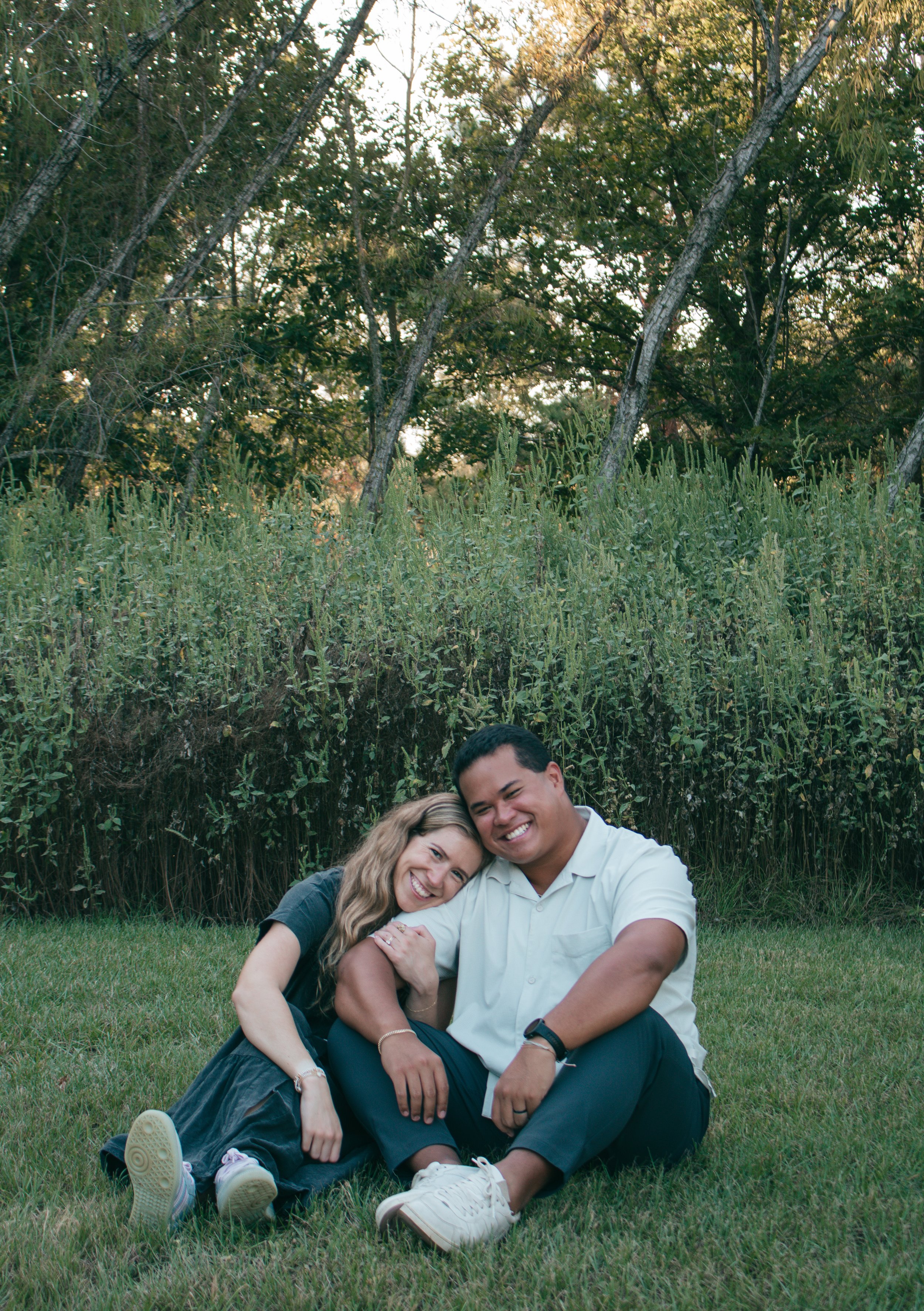 Candid couple’s photo in nature: one leans on the other’s shoulder as they smile, framed by greenery and soft light.