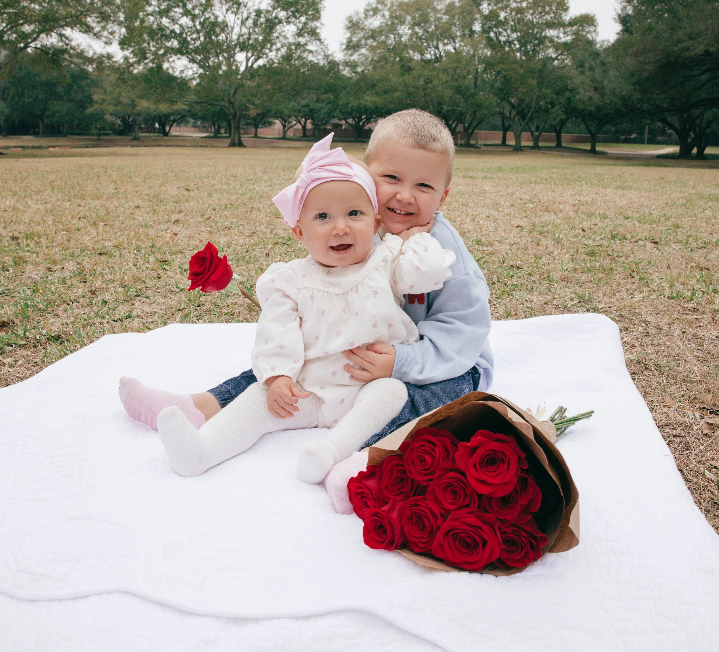 Portrait of siblings in a serene park, seated on a white blanket with red roses and soft greenery in the background.