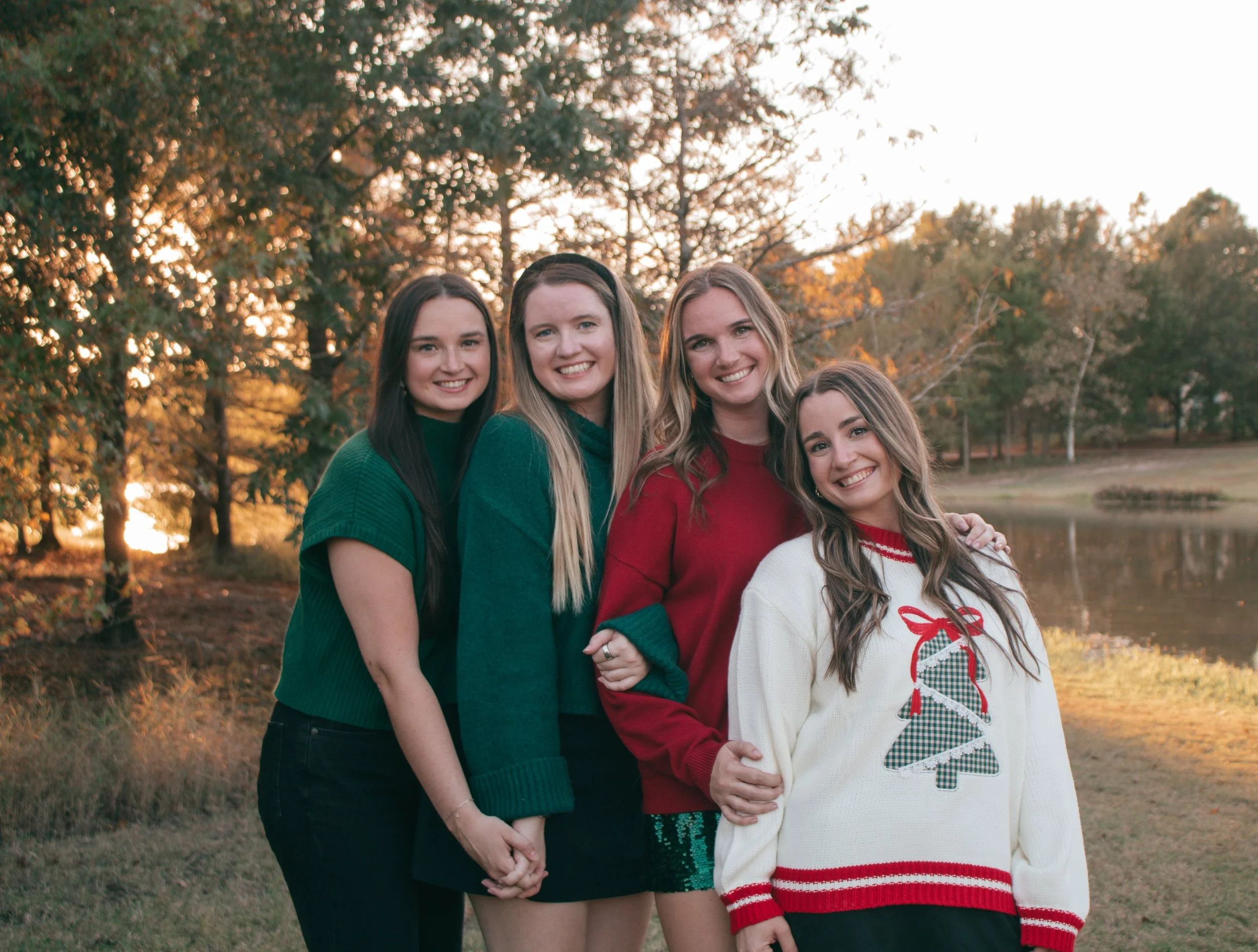 Group portrait of four sisters in festive sweaters, gathered near a wooded pond in soft evening light.
