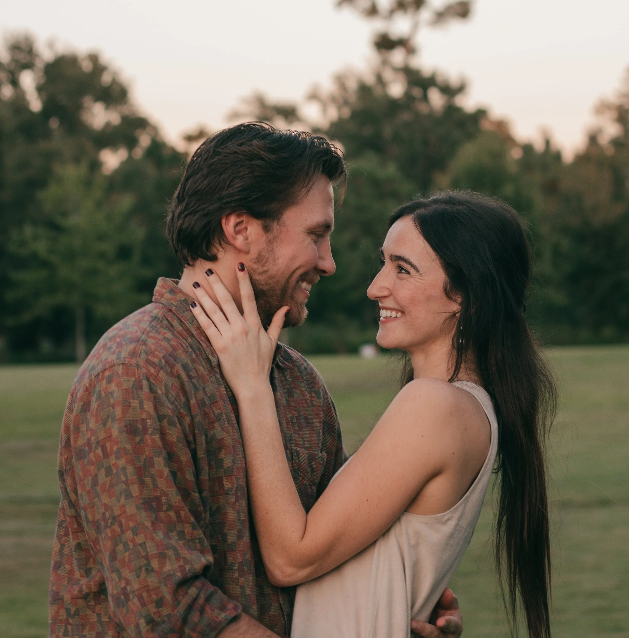 Romantic outdoor portrait of a smiling couple in warm light, sharing a sweet moment in a grassy, tree-lined setting.
