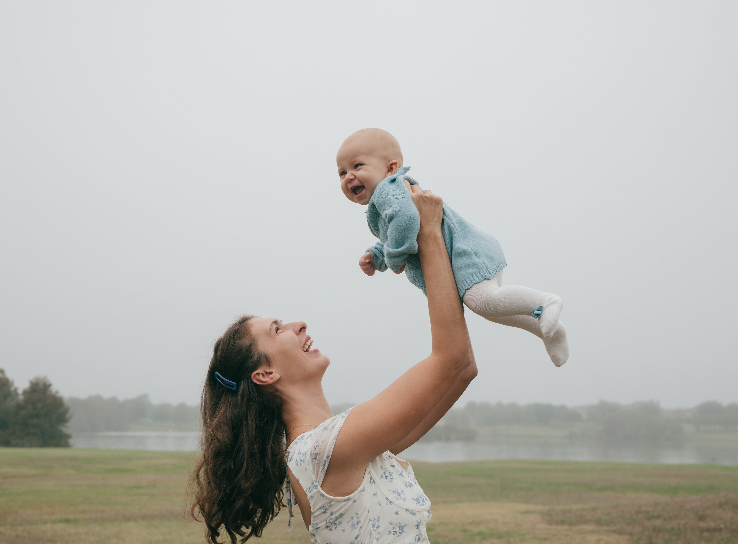 Mom and baby share a playful moment outdoors, baby smiling as mom lifts her into the air.