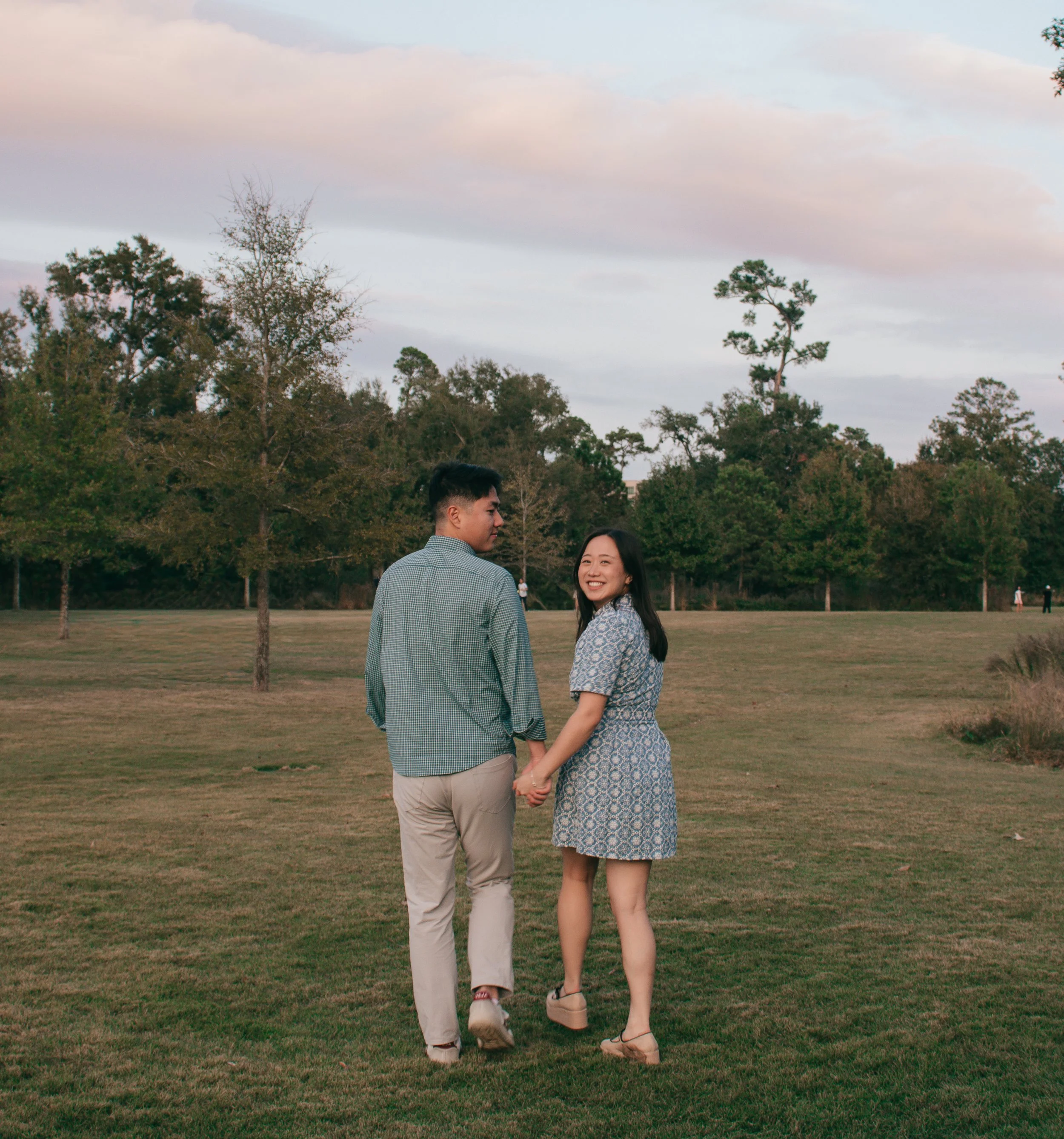 Natural photo of couple in a serene field, one turning back with a joyful smile as they walk beneath a pink‑hued sky.