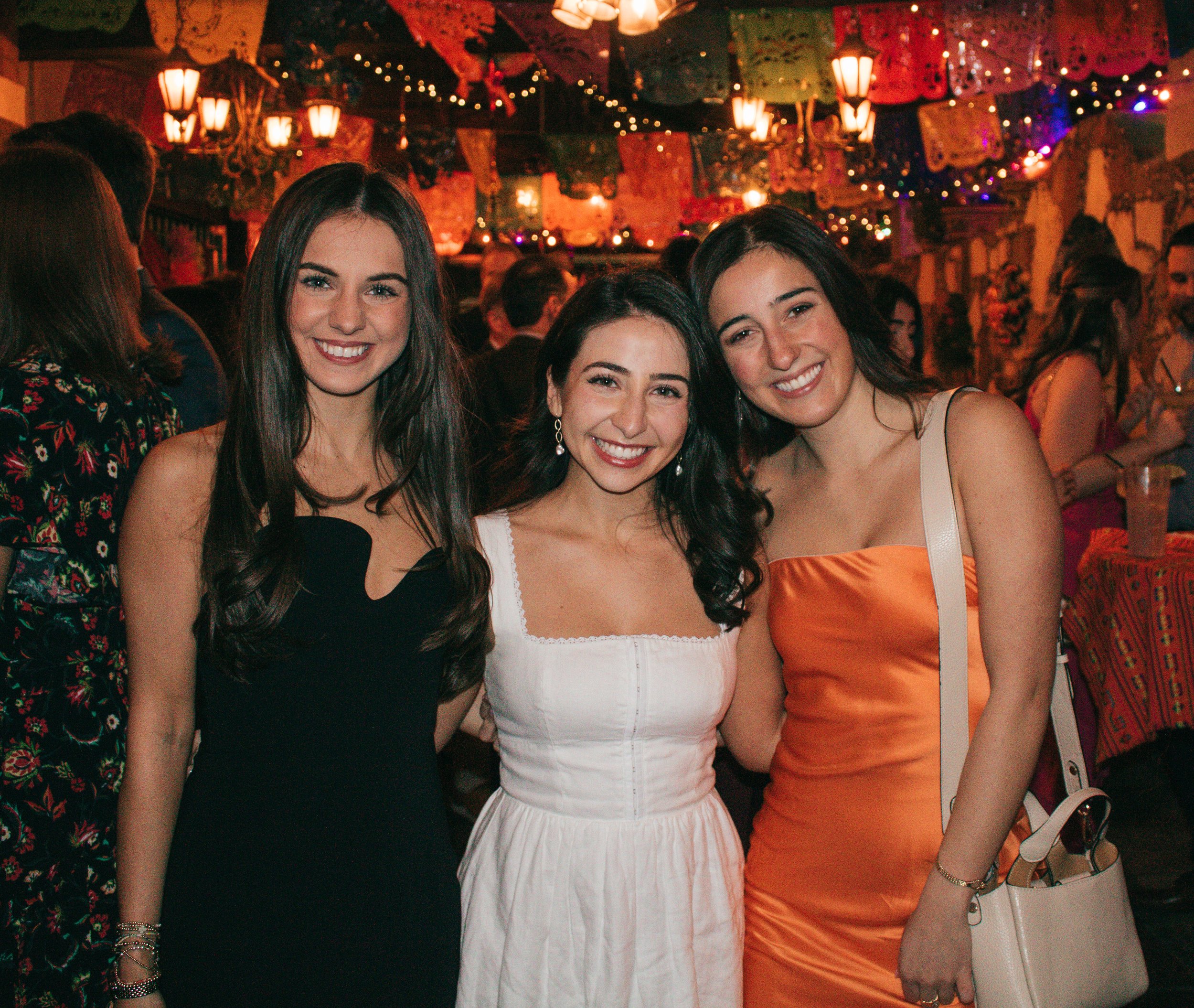 Trio of guests in formal attire pose at a lively pre-wedding event with festive lights and colorful decorations.