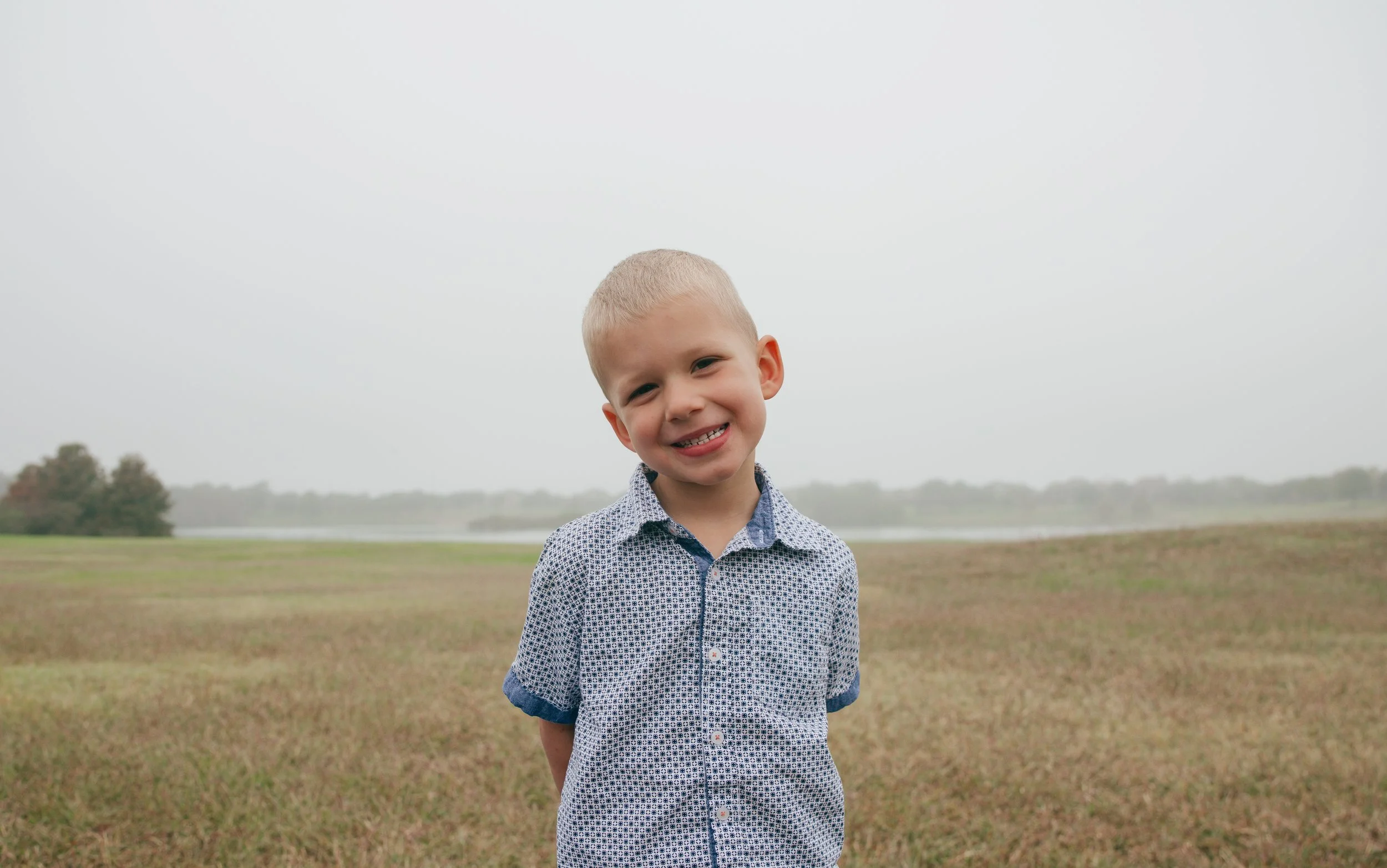Smiling boy standing outdoors, surrounded by greenery and soft natural light in a peaceful nature setting.