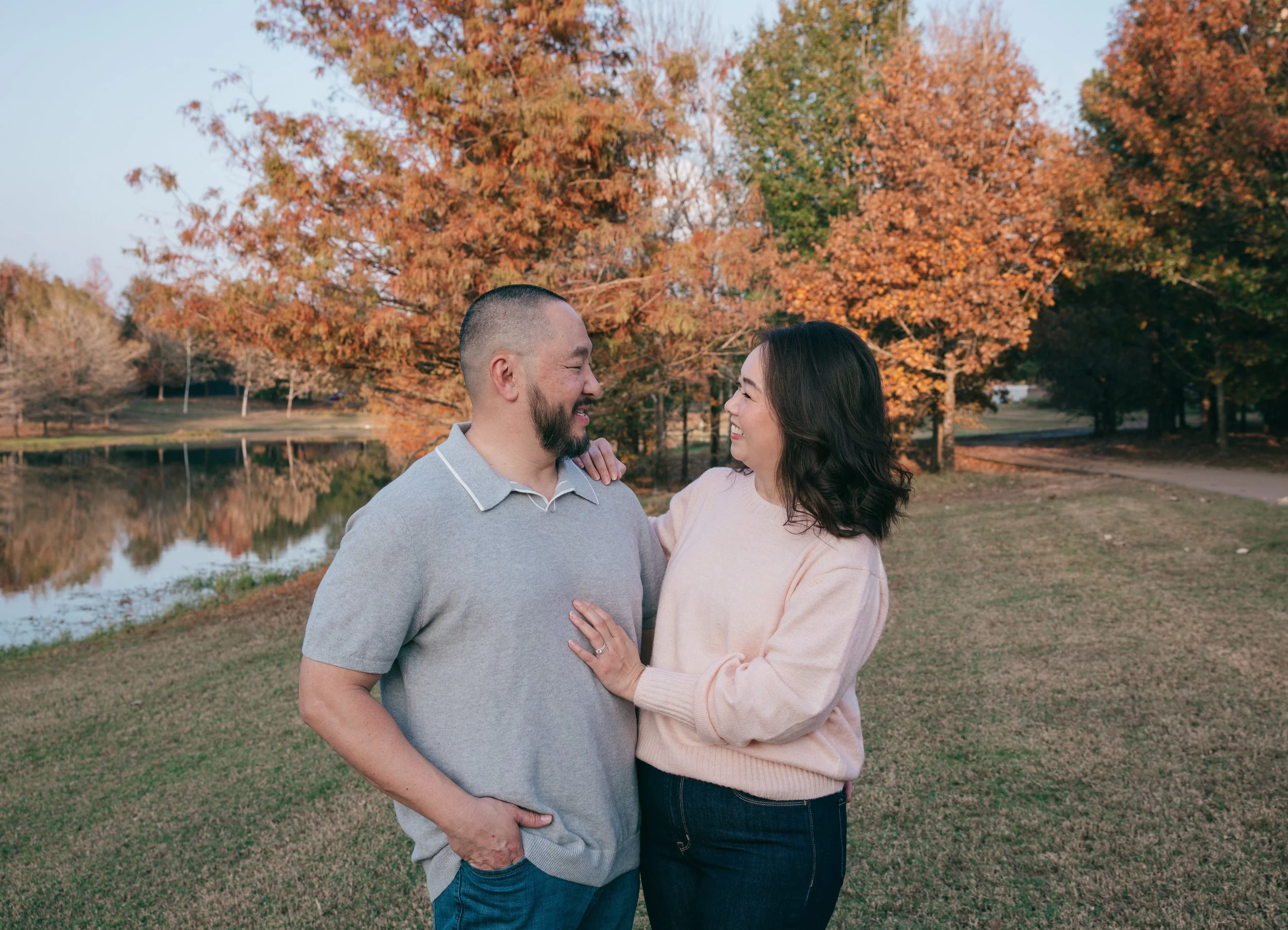 Outdoor portrait of couple facing each other, smiling with hands touching, set against a fall-colored lakeside view.