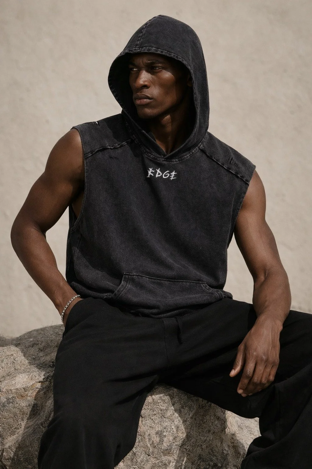 A young man with dark skin tone wearing a black sleeveless hoodie with the hood up, black pants, and a silver bracelet, sitting on a rock against a plain beige background.