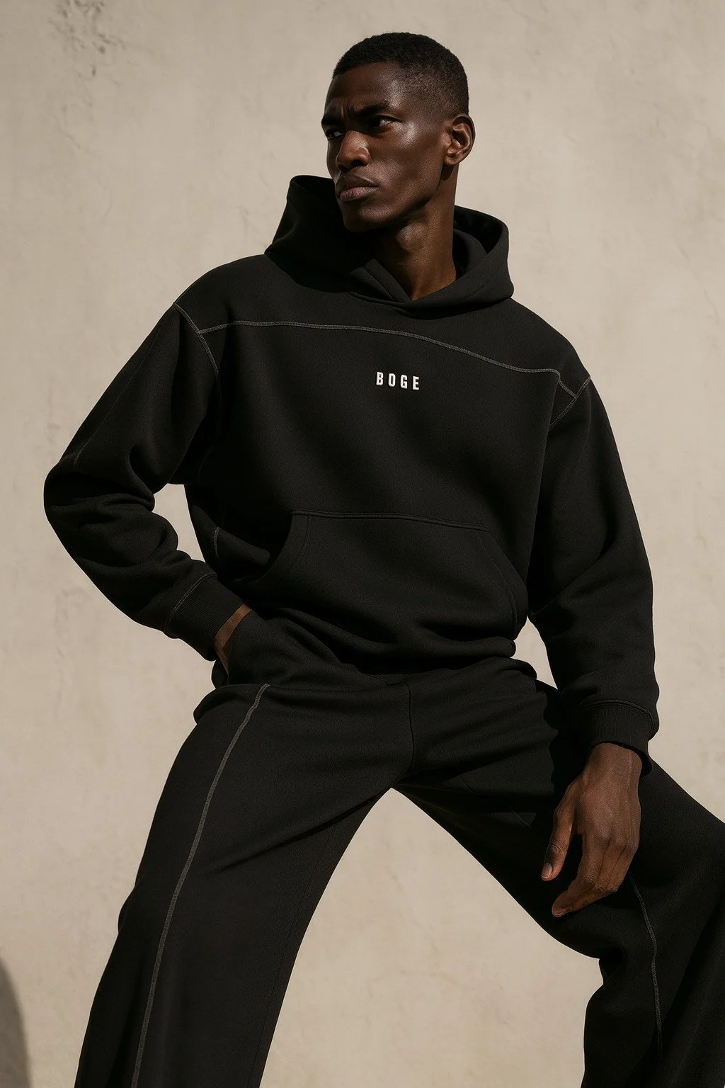 A young man with dark skin and short hair wearing a black hoodie and black pants, standing against a plain light-colored background.