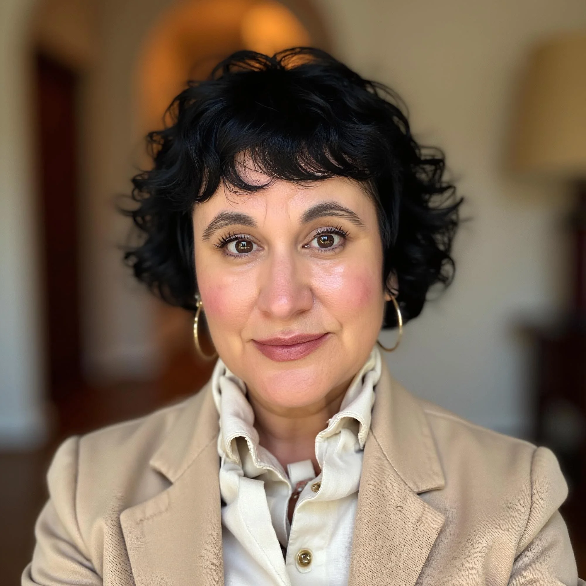 A woman with short, curly black hair wearing a beige blazer and a cream-colored blouse, smiling indoors.