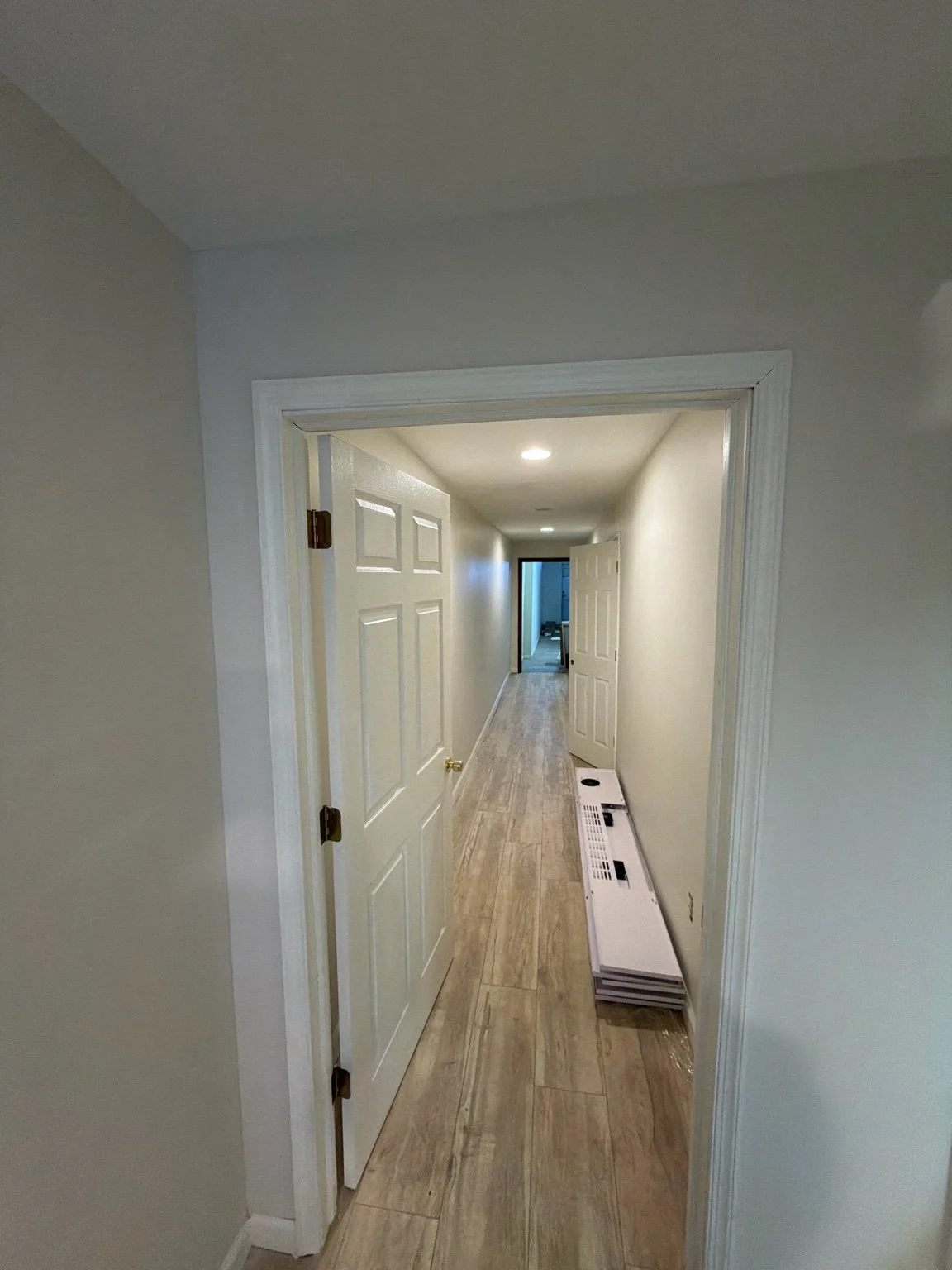 Long hallway with white walls, wooden flooring, and a door slightly ajar on the left side. There is a white radiator cover on the right, and recessed lighting in the ceiling. At the end of the hallway, a room with blue lighting is visible.