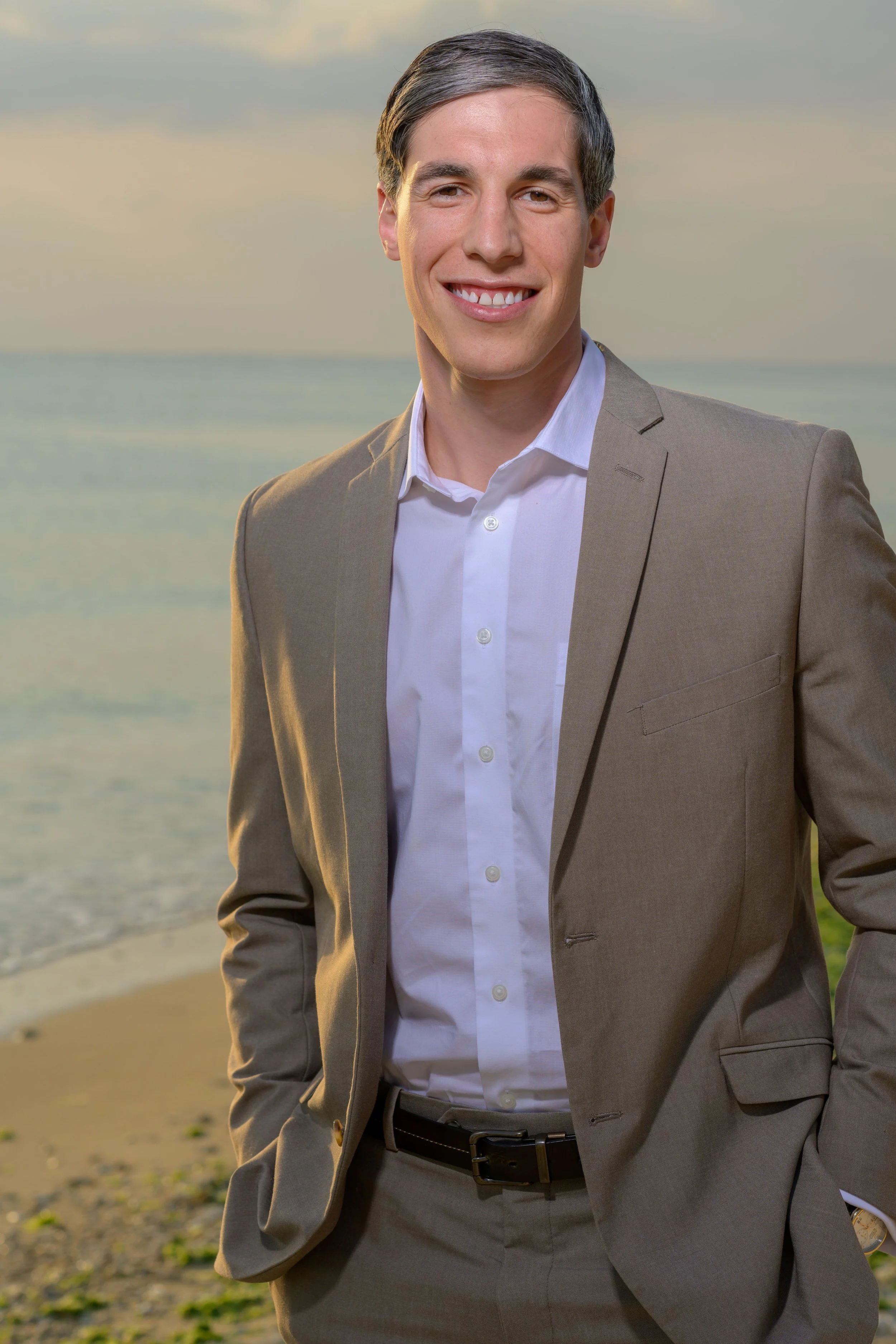 A young man in a beige suit and white shirt smiling at the camera on a beach at sunset.