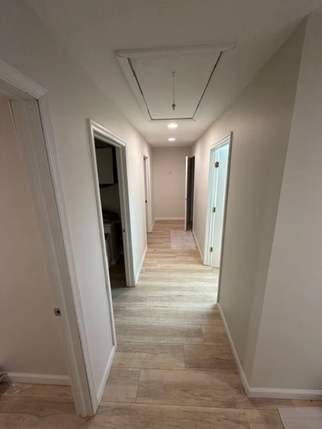 Empty hallway in a house with white walls, light-colored wood flooring, multiple doorways, and an unfinished ceiling with a hanging light fixture