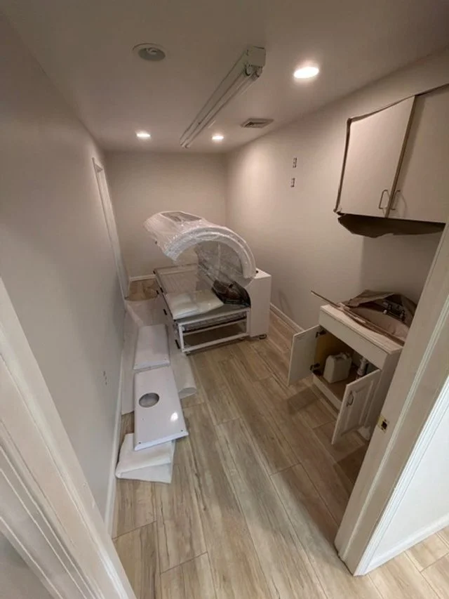 A small room with a portable CT scan machine, an open cabinet, a small table, and some rolled-up items on the floor.