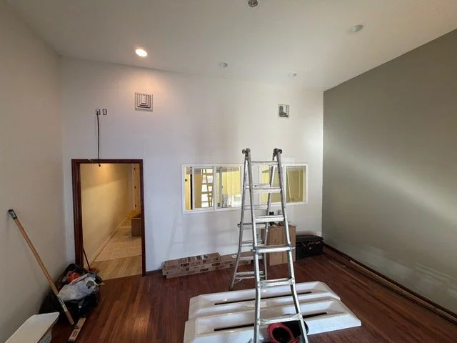 A room under renovation with a step ladder, construction materials, and tools on wooden flooring, a small window opening in the wall, two ceiling vents, and a doorway leading to another room.