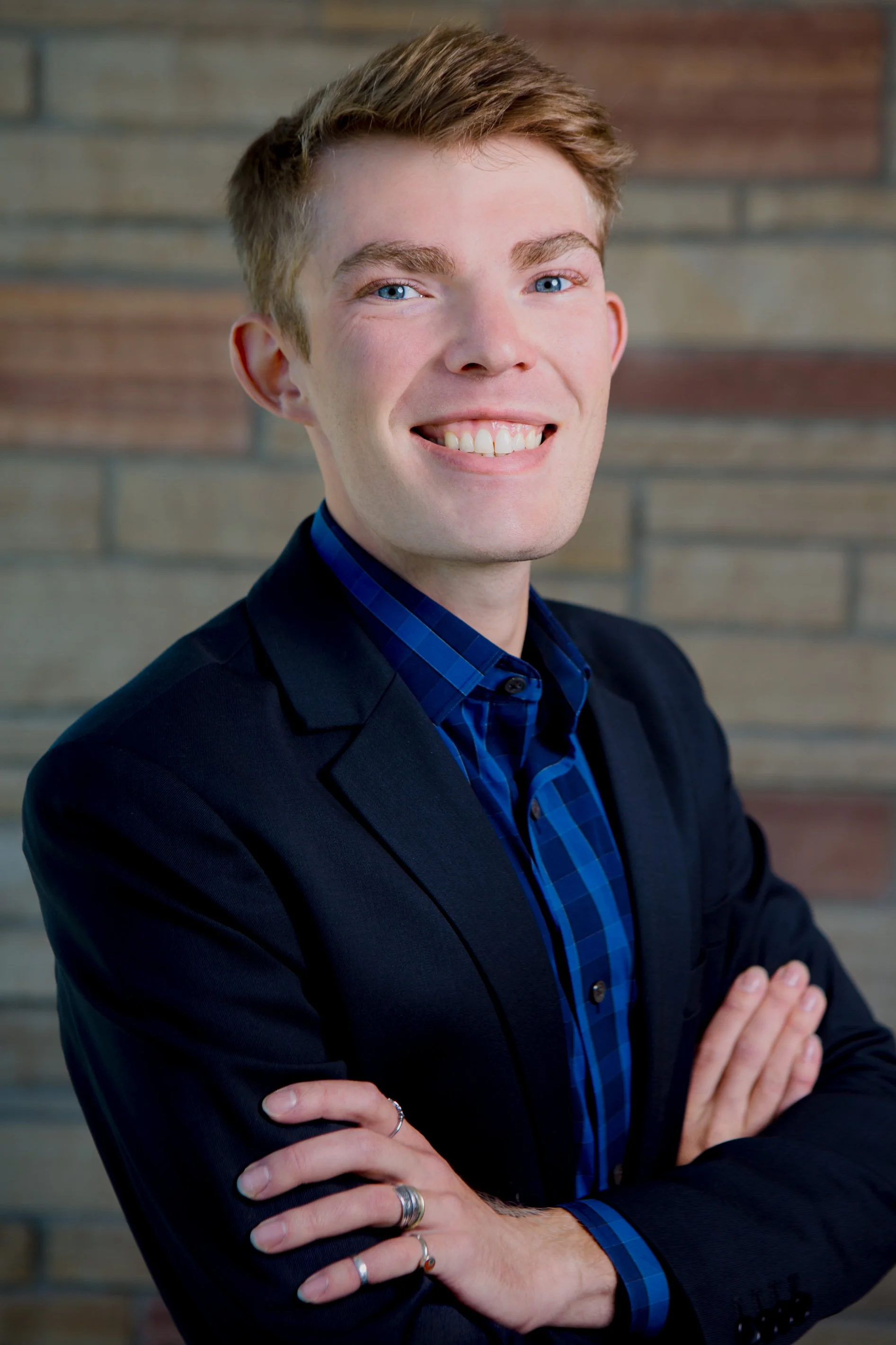 A smiling young man with light brown hair, blue eyes, wearing a black blazer over a blue plaid shirt, standing with arms crossed in front of a brick wall background.