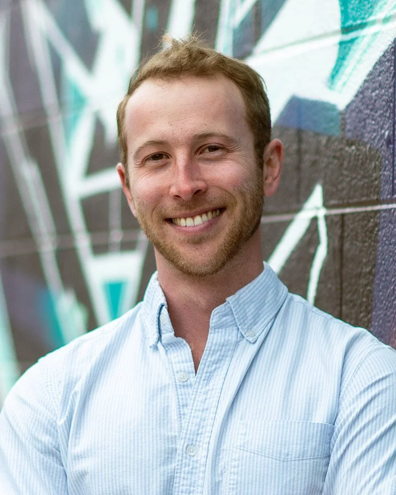 A smiling man with a light beard and short, light brown hair standing against a graffiti wall.
