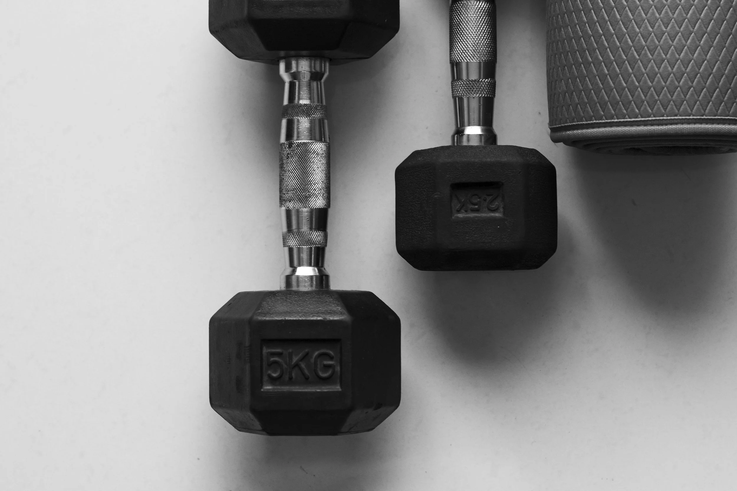 Two black 5-kilogram dumbbells with metal handles and textured grips lying flat on a plain white surface with a textured foam roller nearby.