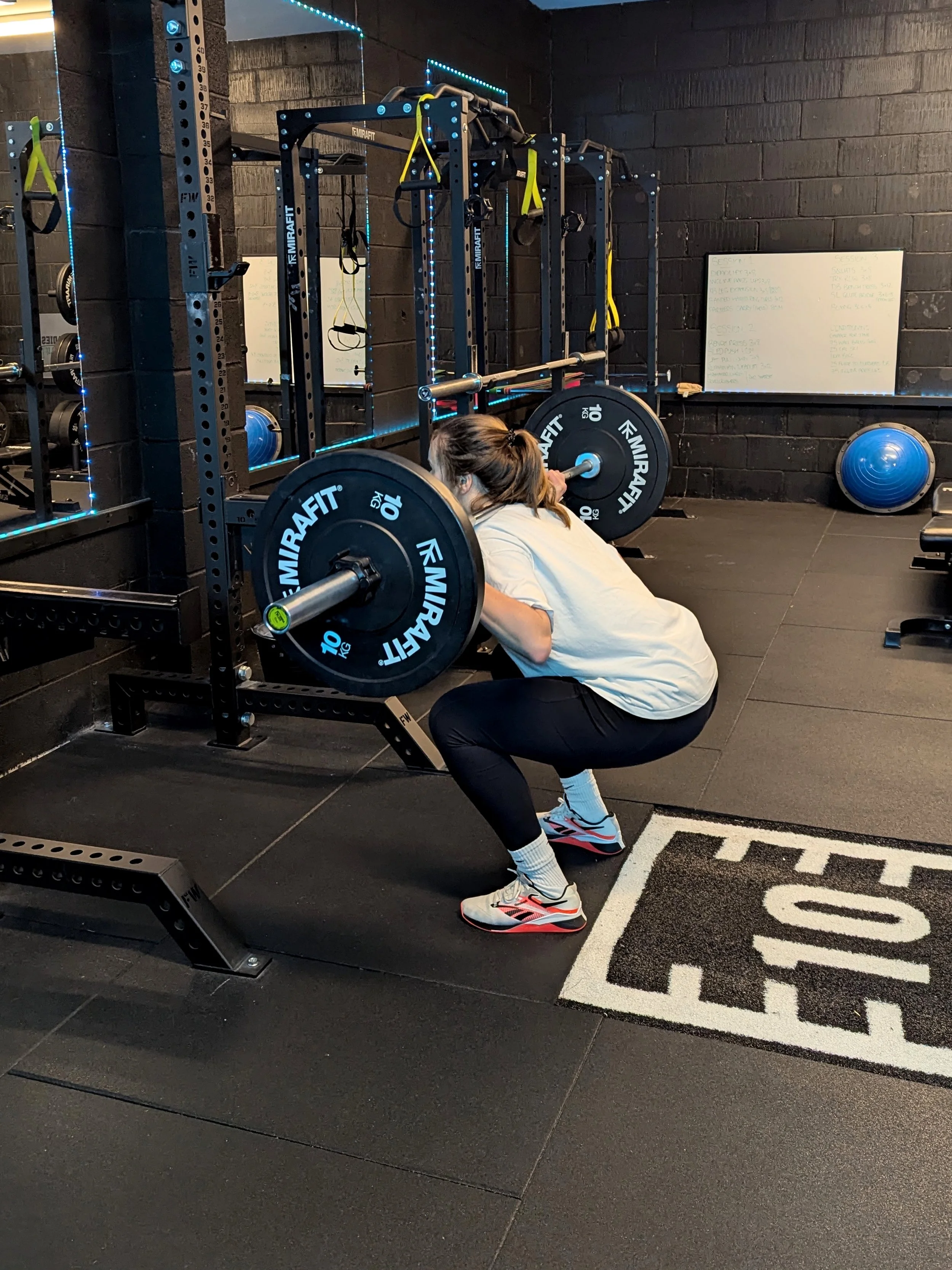 A woman performing a squat with a barbell loaded with weights in a gym, facing a black wall with a whiteboard and equipment around.