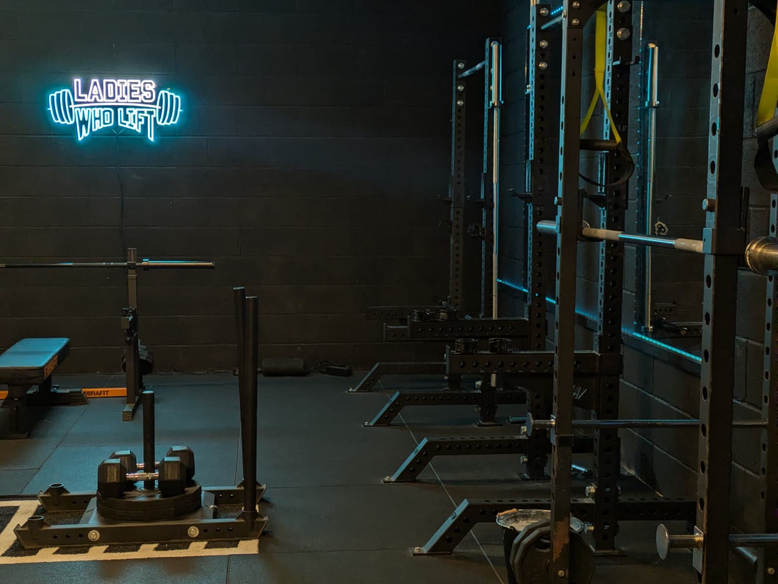 A dimly lit gym with black walls and floor, featuring a neon sign that says 'Ladies Who Lift' with a barbell icon on the wall. There is a weightlifting bench on the left, a weight sled with weights in the foreground, and a large black metal power rack with various attachments and weights on the right.