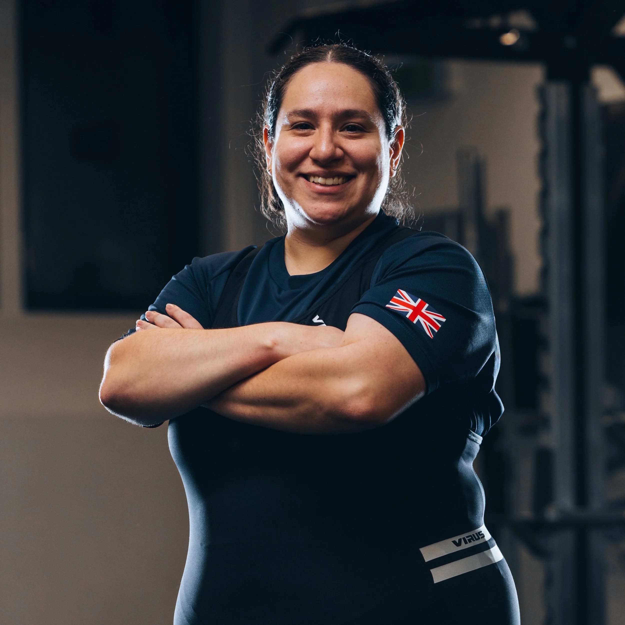 A smiling woman with arms crossed, wearing a black sports shirt with a UK flag patch on the sleeve, standing in a gym.
