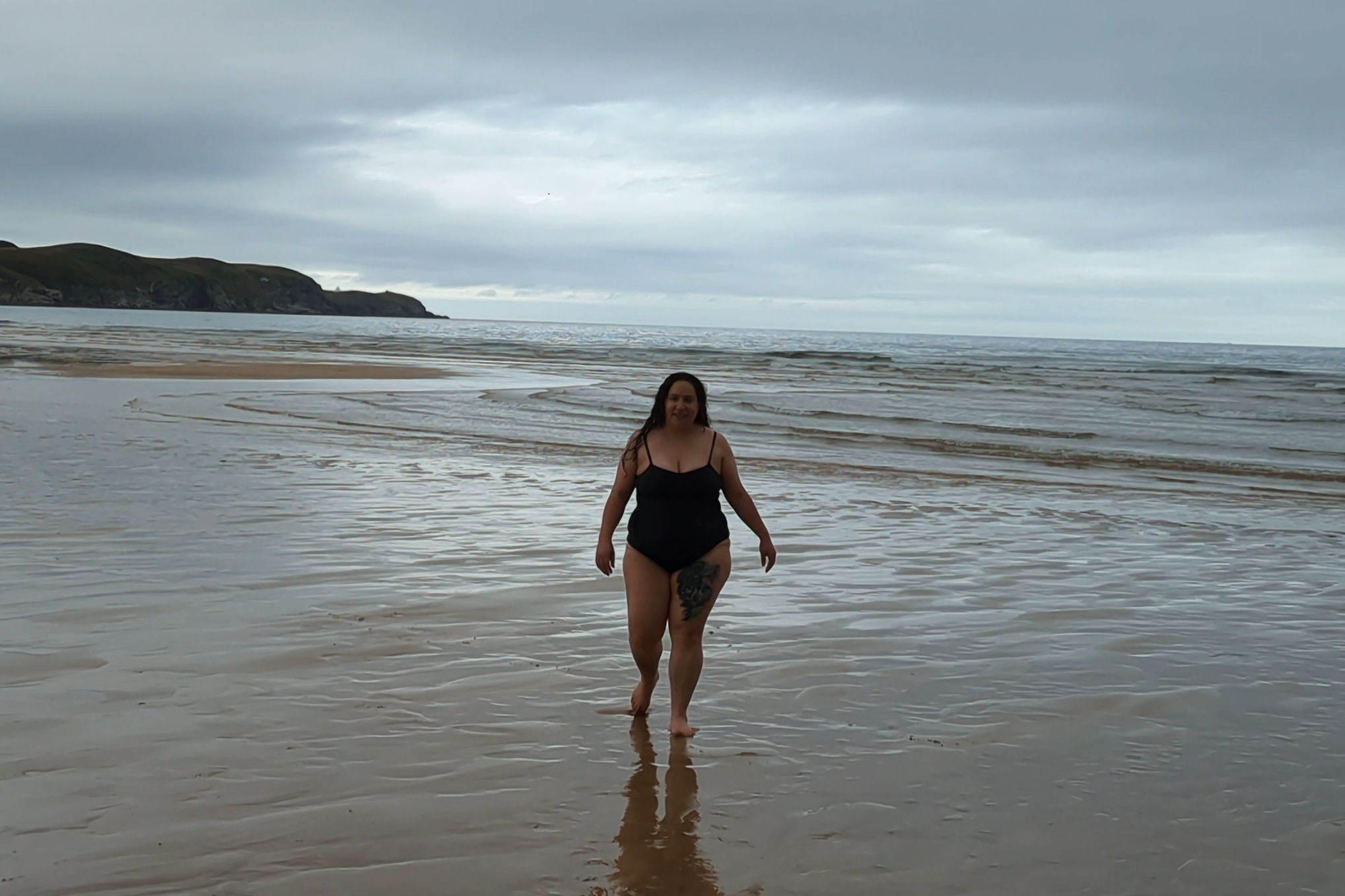 A woman walking barefoot on a wet sandy beach with waves in the background. She is wearing a black swimsuit and has a large tattoo on her left thigh. The sky is overcast with gray clouds.