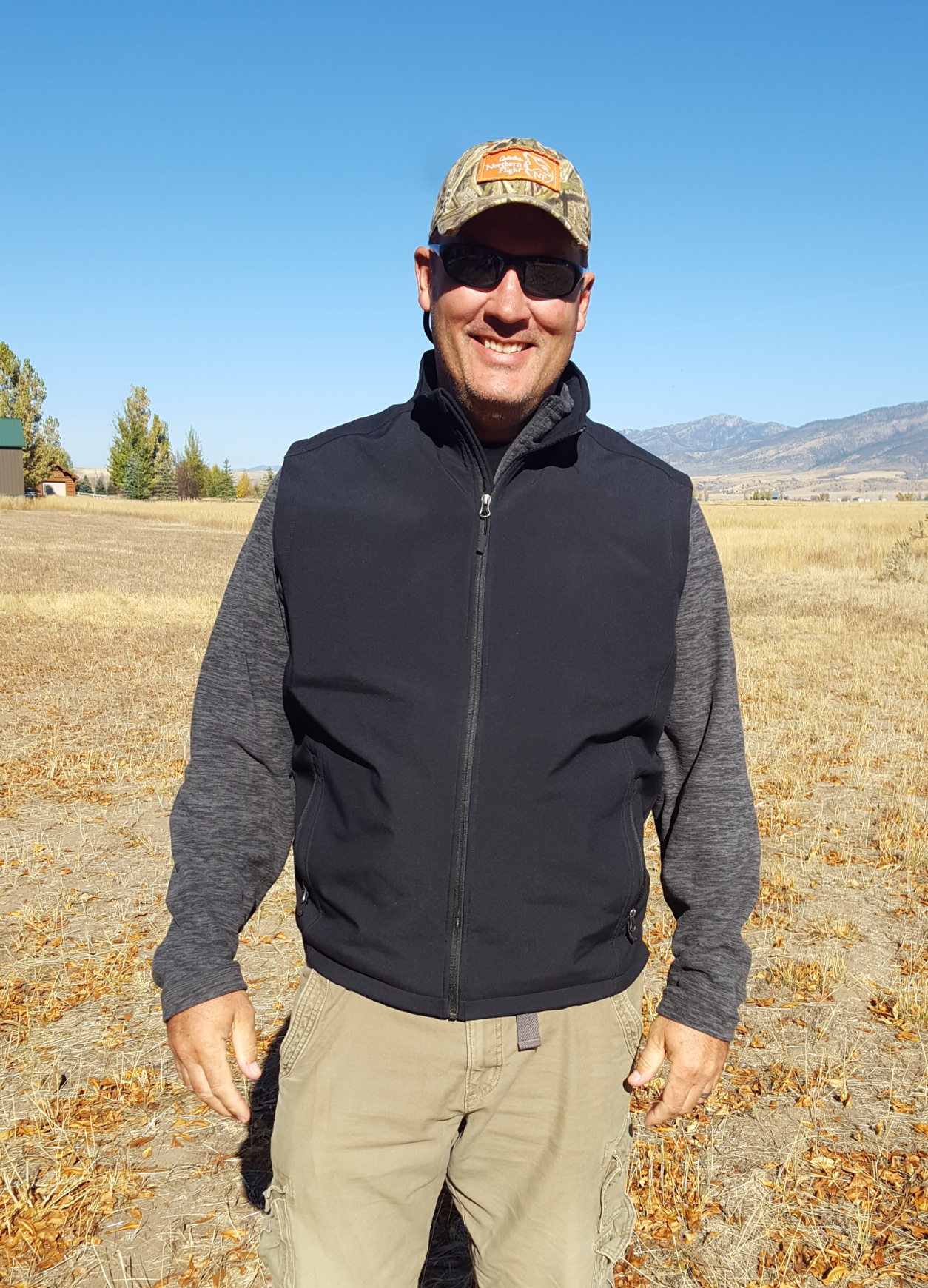 A man smiling outdoors, wearing sunglasses, a camouflage cap, a black vest over a gray and black long-sleeve shirt, and khaki pants, standing in a field with mountains in the background under a clear blue sky.